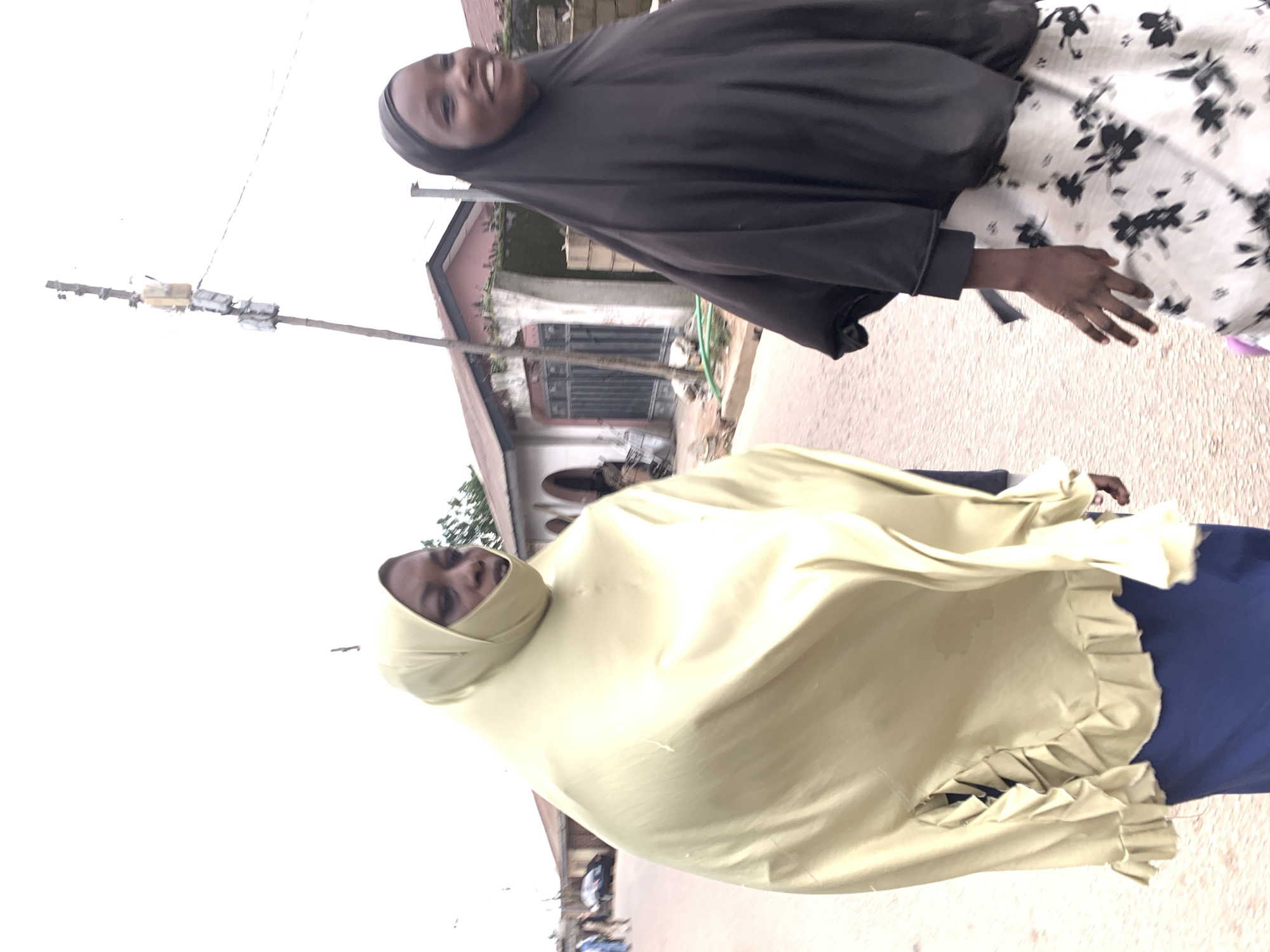 Two individuals are walking on a dirt road in Jos, Nigeria. The person on the left is wearing a light yellow hijab and a blue skirt. The person on the right is wearing a black hijab and a black and white floral skirt. In the background, there are buildings, a utility pole with electrical equipment, and a car. The sky is overcast.