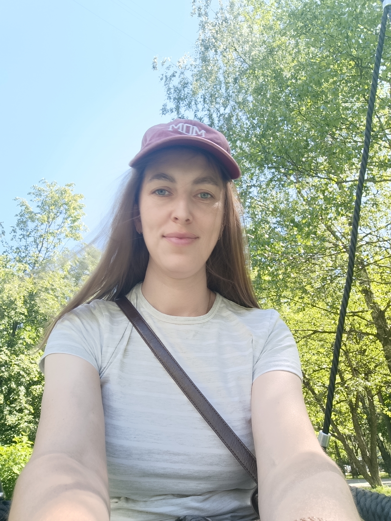 A low-angle self-portrait features a Caucasian female in her 20s or 30s, positioned centrally. She has long, light-brown hair, light-colored eyes, and a faint smile. She wears a maroon baseball cap with "MOM" embroidered in white capital letters on the front, and a light-colored, possibly grey or white, short-sleeved t-shirt with subtle horizontal stripes. A dark brown strap, consistent with a shoulder bag, crosses her torso diagonally from her upper left to her lower right. Her forearms are visible in the lower foreground. The background consists of a clear blue sky on the upper left and dense green foliage from deciduous trees under bright sunlight, indicative of a park or natural outdoor setting. A thick, dark-colored rope with a silver metallic clamp is prominently visible on the right side of the frame, extending vertically. The image contains no discernible landmarks or geographical indicators to confirm a specific location within Moscow, Russia.
