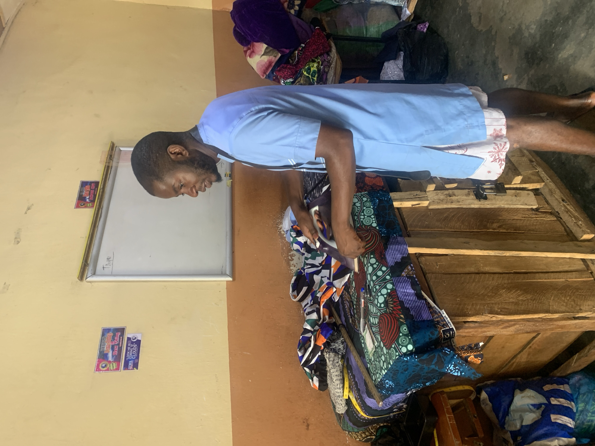 A man wearing a light blue scrub top and patterned shorts stands facing a collection of colorful fabrics. He is looking down and appears to be in the process of selecting or arranging the materials. The fabrics are piled on a wooden surface, possibly a table or shelves, with some extending over the edge. Behind him, more fabrics and items are visible, creating a cluttered but functional atmosphere.
On the wall to his left, there is a whiteboard with some writing on it and two small stickers. One sticker features vibrant colors and text that is difficult to read clearly, but appears to be a promotional or informational label. The other sticker has the text "I LOOK HE IS COMING" and "CLOUDS." The room has a simple, utilitarian feel with light-colored walls and wooden elements. The lighting suggests it is daytime. The scene appears to be in a workshop or shop where fabrics are sold or used for tailoring. The man's focused expression and the organized piles of fabric indicate he is engaged in his work. The overall impression is one of a bustling environment dedicated to textile crafts.