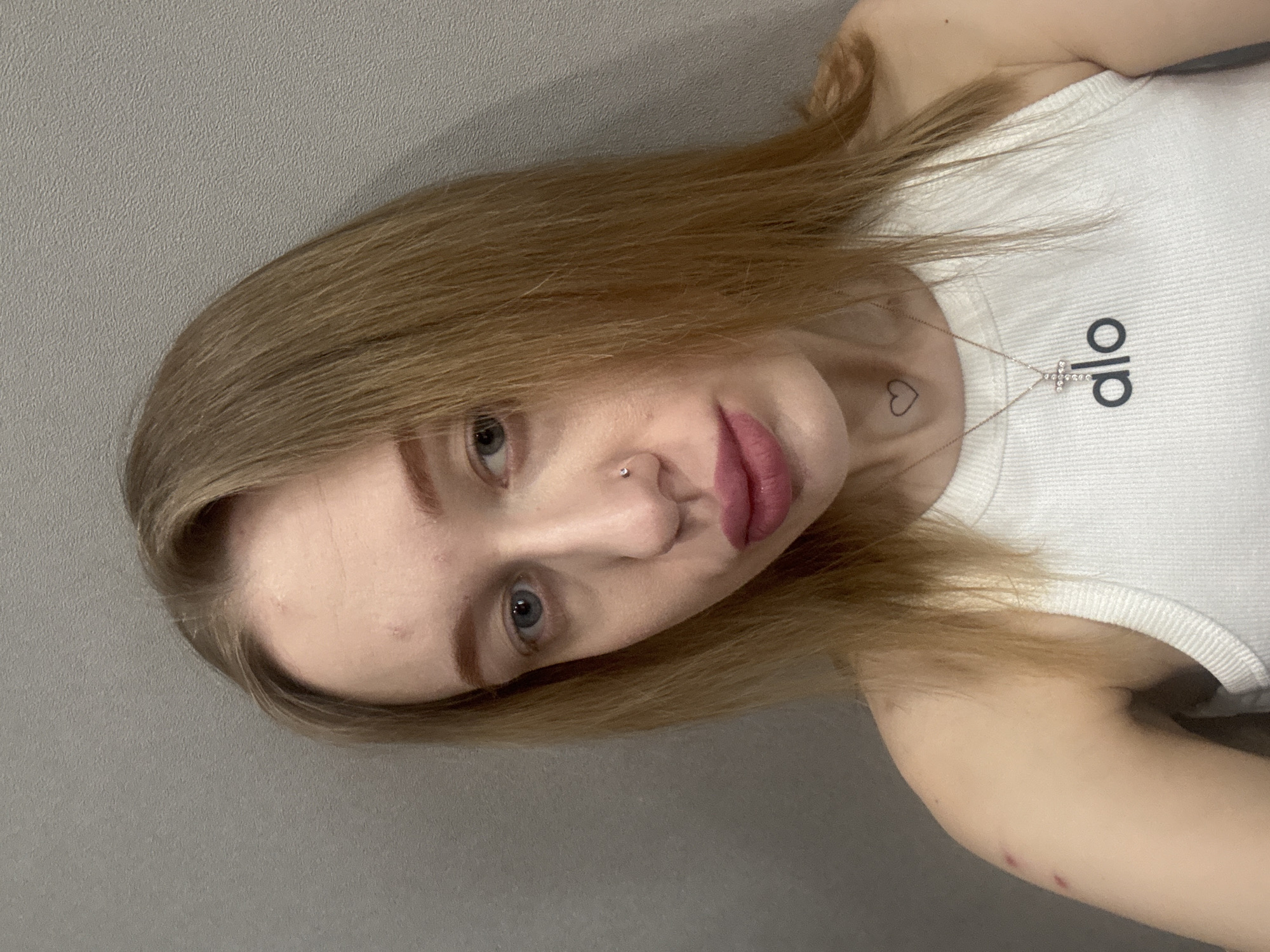 A woman with light brown hair and blue eyes is pictured close-up against a gray wall. She is wearing a white tank top with the word "Alo" printed on it. She has a nose piercing and a small heart tattoo on her neck, as well as a necklace with a cross pendant. There is a mark on her arm. The location in the city of Ovoshchnoy, Russia is not discernible from the provided image.