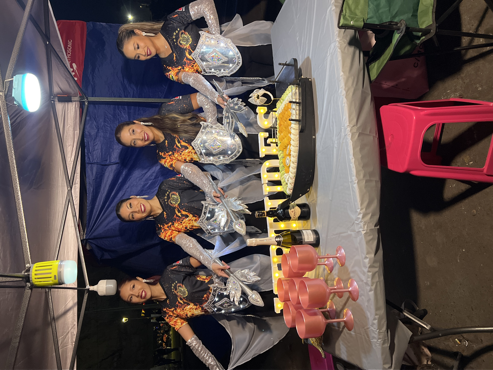 Four women are posing behind a table that is draped with a gray tablecloth. The women are wearing matching black and orange flame-patterned shirts with silver sequined sleeves. They are holding decorative wands that resemble swords. The table is adorned with illuminated letters that spell out "CEFIRO," a row of food items (likely sushi), two wine bottles, and a stack of pink-colored stemware.
The background includes a pop-up tent structure with a blue interior. A light fixture hangs from the tent frame. A red banner with the word "METROSONIC" is partially visible within the tent. Outside the tent, one can see a green folding chair, a pink plastic stool, and a dark, possibly asphalt ground surface. The setting appears to be outdoors at night.
Given the name "CEFIRO" and the location hint (Arica, Chile), it is plausible that this scene is associated with a local event or celebration in Arica.