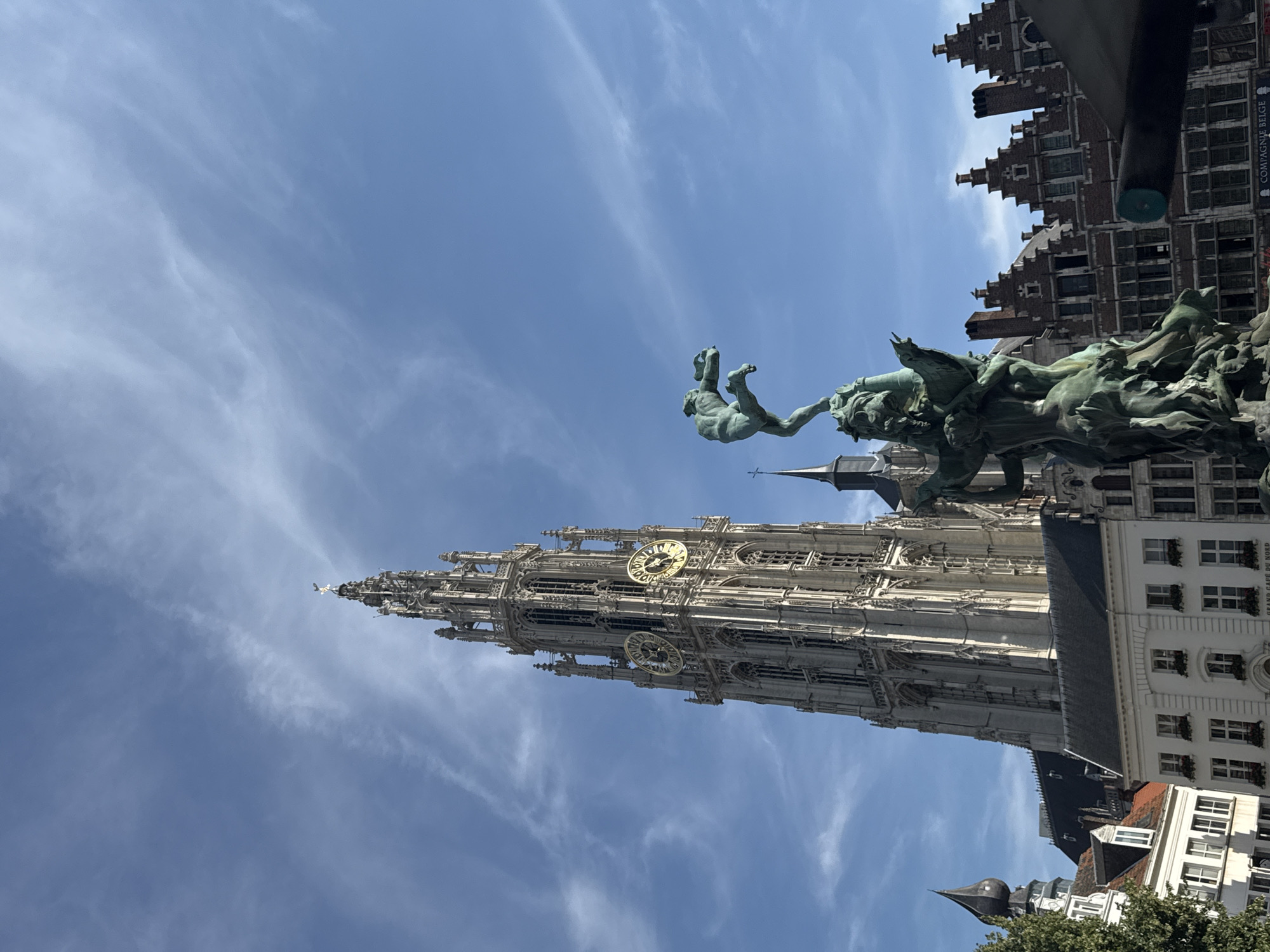 The image shows a low-angle view of the Cathedral of Our Lady in Antwerp, Belgium, with the Brabo Fountain in the foreground. The cathedral's tall, Gothic spire dominates the upper left of the frame, featuring ornate architectural details and clock faces. The Brabo Fountain, a bronze sculpture, is positioned in the lower right, depicting a mythological scene with figures and a dragon-like creature. Behind the fountain, traditional Antwerp architecture with stepped gables is visible. The sky is blue with wispy clouds.