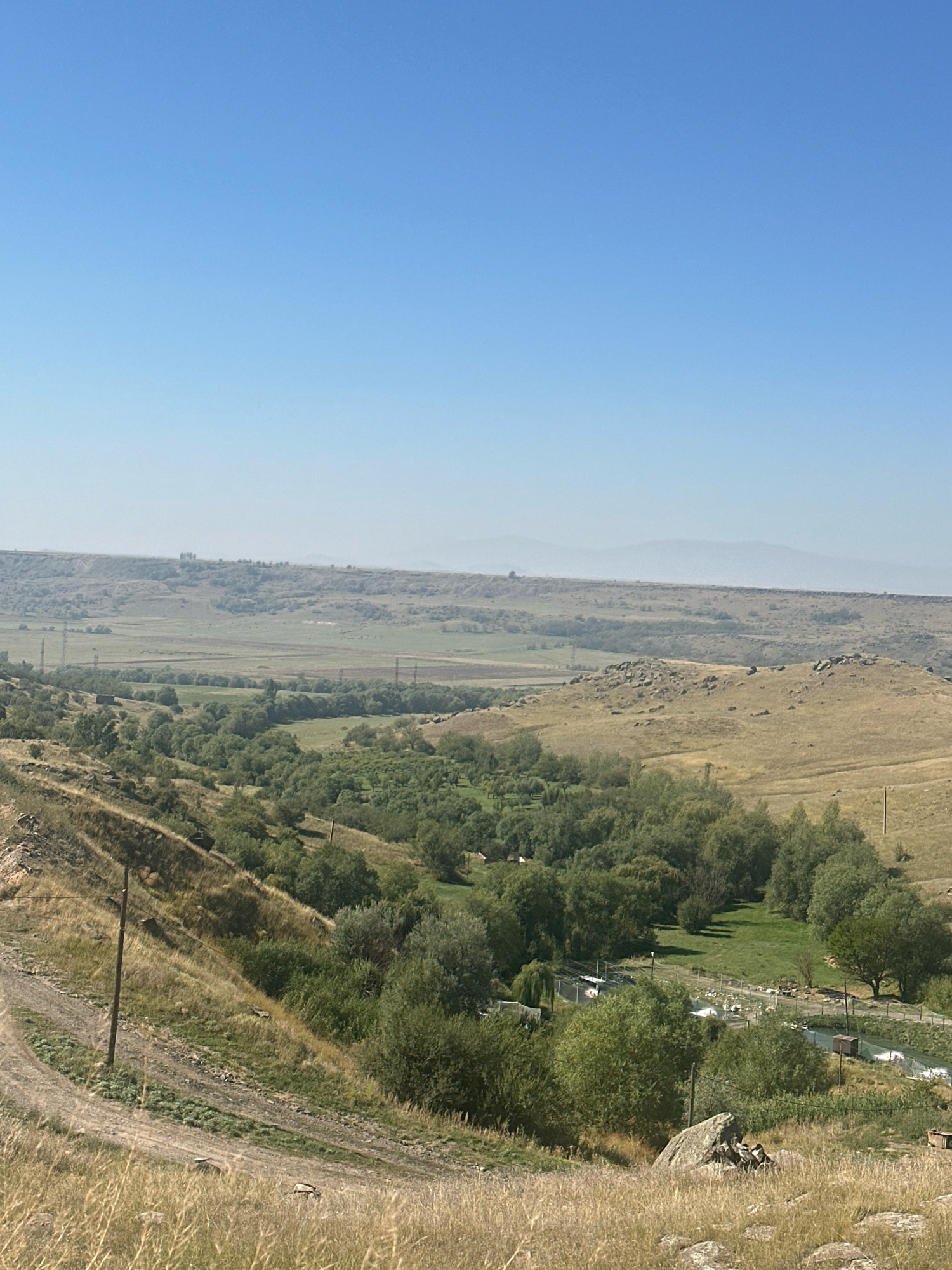 The image depicts a vast, open landscape under a clear blue sky. In the foreground, dry, golden grass covers a sloping hillside, with a dirt road winding its way down. Scattered rocks are visible among the grass.
Further down the slope, a dense cluster of green trees creates a verdant valley. Within this valley, small structures and what appear to be enclosures are nestled among the trees, possibly indicating agricultural activity or a small settlement. Power lines and tall poles traverse the mid-ground, extending across a flatter, cultivated area.
In the background, rolling hills stretch towards the horizon, gradually fading into a hazy, muted landscape under the vast expanse of the sky. The overall impression is one of rural tranquility and expansive natural beauty, with hints of human presence in the form of infrastructure and possibly small dwellings. While the exact location within Gyumri, Armenia cannot be definitively determined from the image alone, the landscape is consistent with the general terrain found in and around the city, which is known for its hilly surroundings and open plains.