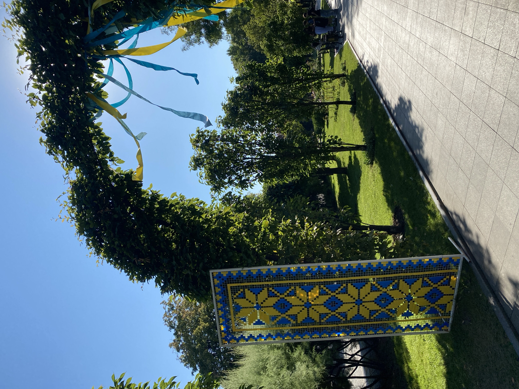 A vertical, rectangular artwork featuring a Ukrainian traditional pattern in blue and yellow is displayed outdoors, leaning against a metal frame. The artwork is positioned on a grassy area next to a paved walkway. Above and behind the artwork, a green archway formed by intertwined branches is decorated with flowing blue and yellow ribbons, which are gently swaying. The sun is visible through the archway, casting rays through the leaves and ribbons. Beyond the archway and walkway, a park-like setting with numerous trees, green grass, and several people walking in the distance is visible under a clear blue sky. The scene appears to be a public space, possibly a park or a promenade, decorated for a patriotic occasion.
