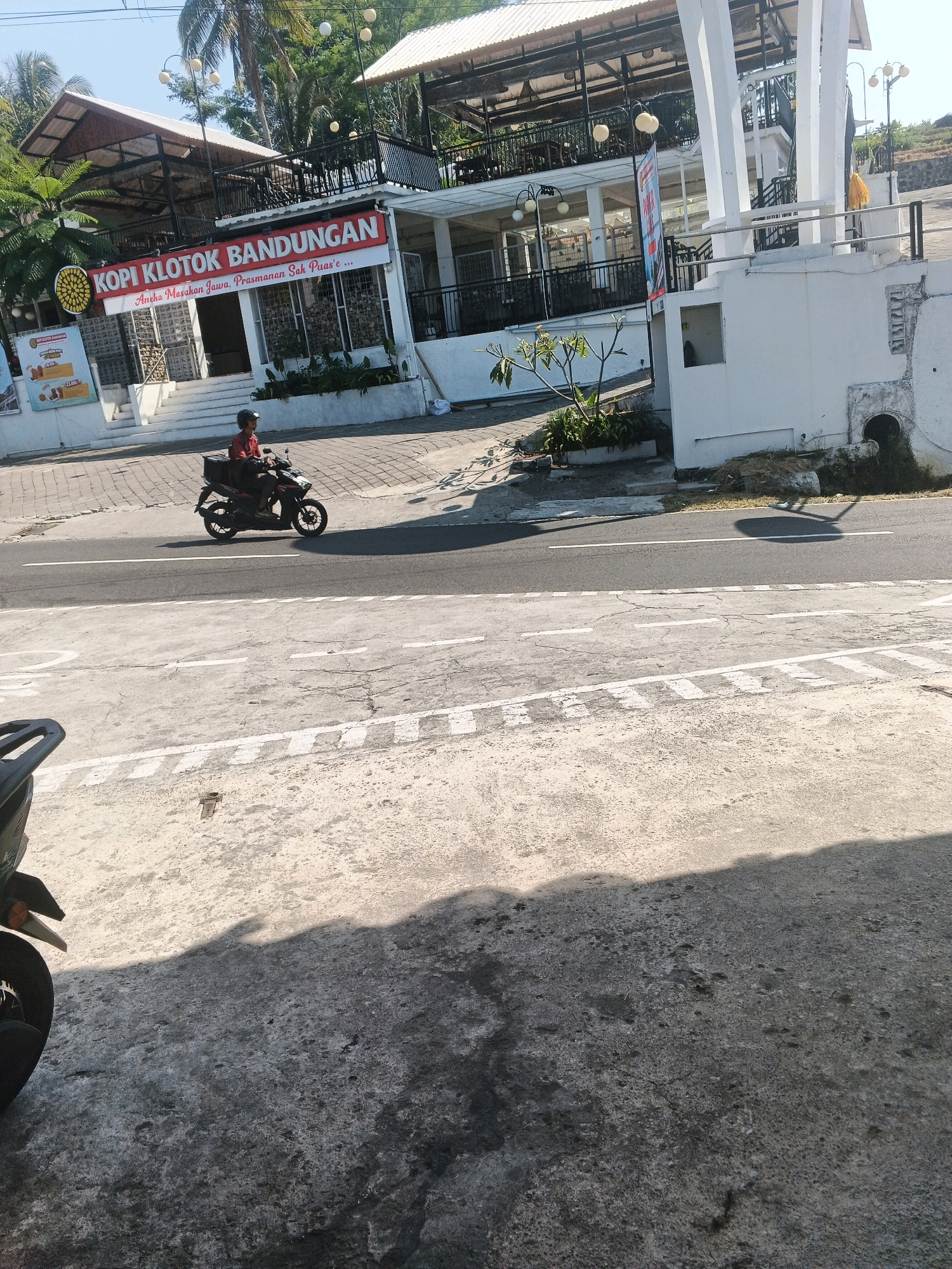 The image depicts a street scene in Pakopen, Indonesia, likely on a sunny day. In the foreground, a person wearing a red shirt and a helmet rides a dark-colored motorcycle with a black storage box on the back. The motorcycle is on a paved road with white lane markings. To the left of the motorcycle, partially visible, is the back of a black motorcycle, suggesting another vehicle is parked or present off-camera.
In the background, a two-story building with a white facade and white columns stands prominently. The upper level appears to be an open-air dining area with tables and chairs, covered by a slanted roof. A red banner with white text reading "KOPI KLOTOK BANDUNGAN" and "Aneka Masakan Jawa, Prasmanan Sak Puas'e..." is displayed on the lower part of the building. Trees, including palm trees, are visible behind and around the building, with some foliage extending into the scene. The sky is clear and blue. The overall environment suggests a commercial area, possibly a restaurant or cafe, situated along a road.