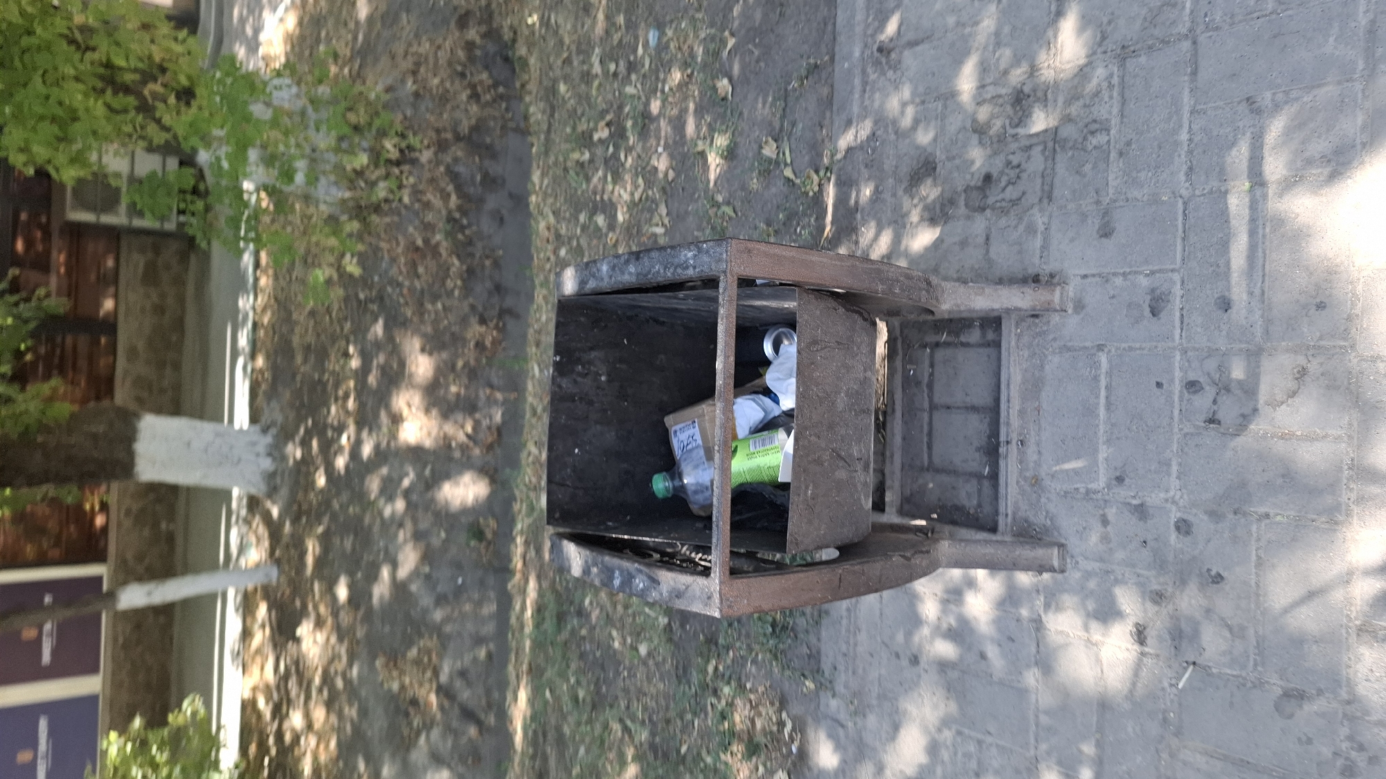 An eye-level, medium shot captures a metal trash receptacle positioned on a paved sidewalk. The receptacle, constructed from dark metal with a rustic appearance, is filled with various discarded items, including a green plastic bottle and a brown cardboard box. In the background, a tree with a white-painted trunk stands in a patch of dirt and dry leaves, with dappled sunlight falling on the ground. Beyond the tree, a low stone wall and the facade of a building are visible, suggesting an urban or semi-urban environment. The sidewalk itself is made of rectangular grey paving stones, with shadows of tree branches cast across its surface. The scene depicts an outdoor setting, likely a public area in Shymkent, Kazakhstan, characterized by the presence of street furniture and landscaping.