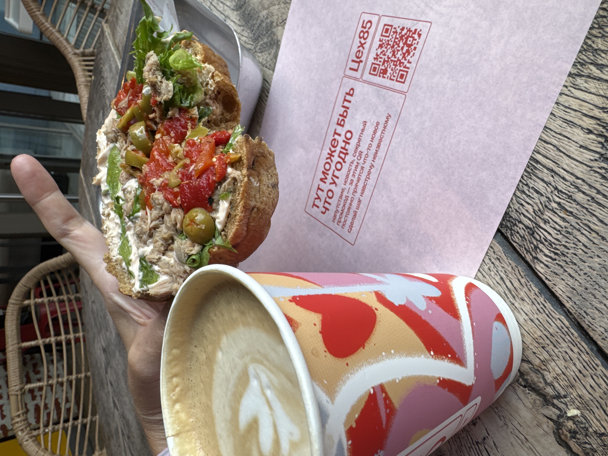 A close-up shot shows a person's hand holding a sandwich over a cup of coffee and a pink piece of paper with Russian text and QR codes. The sandwich is open-faced, with ingredients like tuna, tomatoes, olives, lettuce, and a creamy sauce visible on a piece of toasted bread. The coffee cup has a decorative pattern of hearts and abstract shapes. The pink paper has a heading in red text that translates to "Here can be anything you want." Below that, smaller text says "A surprise, a secret news, a promo code - for this QR. The promo code inspires something new. Take a step towards the unknown." The text is accompanied by two red QR codes. The background shows a wooden table and a wicker chair, suggesting an outdoor cafe setting. This scene is likely from Saint Petersburg, Russia, as indicated by the Russian language text.