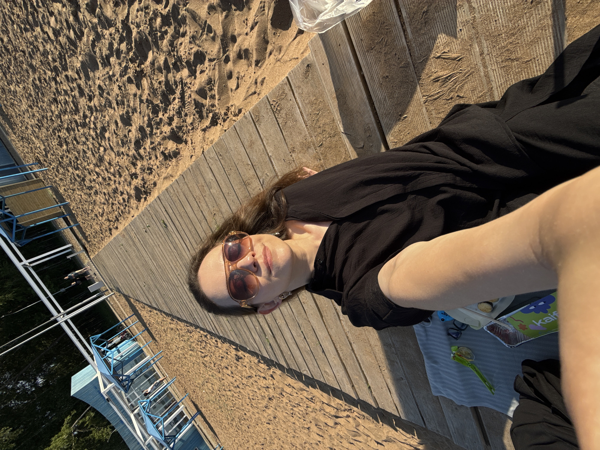 A woman is taking a selfie while lying on a wooden boardwalk next to a sandy beach in Chaikovsky, Russia. She is wearing sunglasses and a black dress, and her arm is extended towards the camera. In front of her, on a blue towel, are a juice box, a small plate with cookies, a green object, and a pair of glasses. In the background, there is a blue building with a white roof and blue canopy structures, some trees, and other people walking on the beach. The sun is casting long shadows across the boardwalk.