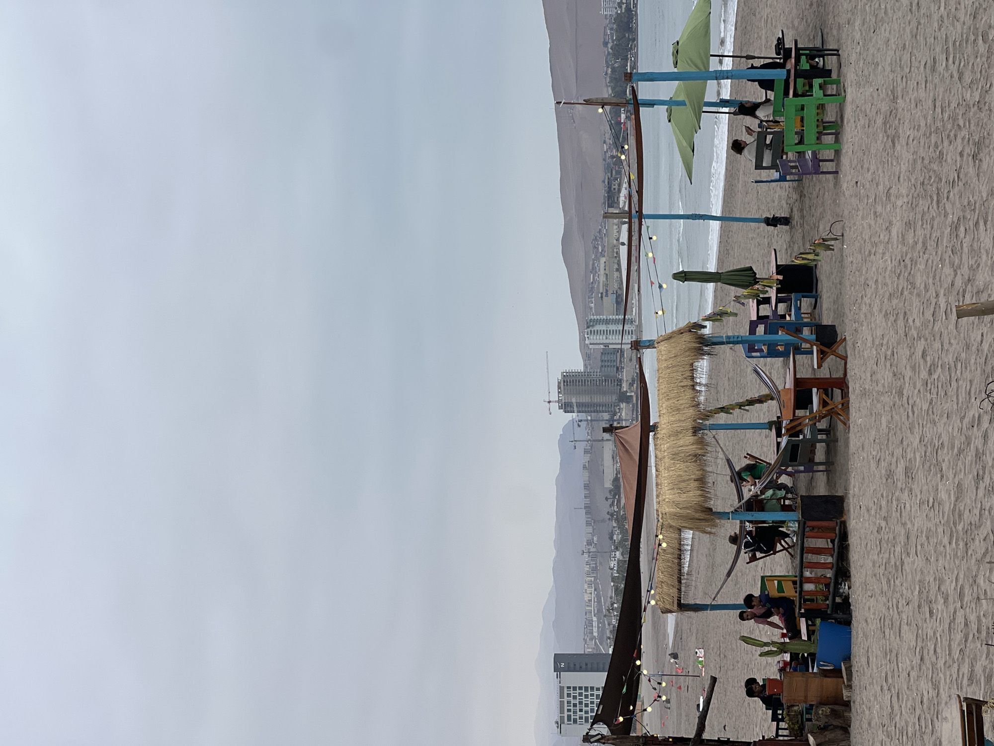 The image shows a beach scene in Arica, Chile. A series of beachside structures, likely cafes or bars, are set up along the sand. These structures feature colorful chairs and tables, straw umbrellas, and decorative lights. People are seen sitting at the tables. In the background, a city skyline is visible, backed by rolling hills or mountains. The sky is overcast. The image is oriented vertically.