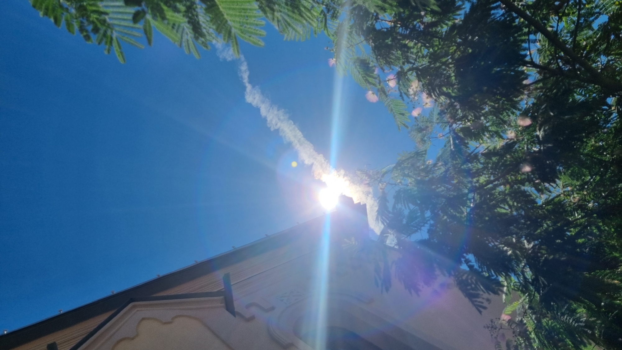 A low-angle, vertical photograph captures a bright, sunny sky over Berehove, Ukraine, with a white contrail arcing diagonally from the lower right to the upper left. The sun is intensely bright, causing lens flare with blue and purple streaks radiating outwards. In the lower left, a portion of a building with decorative trim is visible, its light-colored facade angled upwards. Lush green foliage from trees frames the right side of the image, with some delicate pink flowers interspersed. The sky is a deep, clear blue.