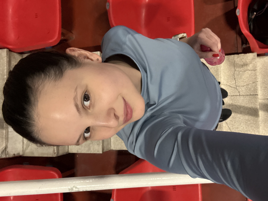 A woman in a light blue long-sleeve shirt is angled towards the camera in a selfie. She has dark hair pulled back, fair skin, and multiple piercings in her ear. Red stadium seats are visible around her. She is holding a pink object in one hand. Her eyes are brown. The background includes gray stairs and dark footwear.