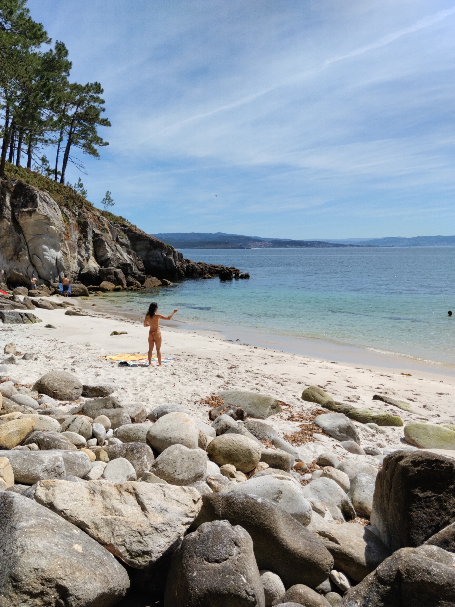 The image depicts a rocky beach on a sunny day. In the foreground, large, grey boulders are scattered across the sand, some covered in moss. A woman in a bikini stands on the sand near the water's edge, facing away from the camera and gesturing with her left hand. Behind her, the sand slopes up to a rocky cliff face with pine trees growing on top. The sea is a clear, turquoise color, with gentle waves lapping at the shore. In the distance, a hazy coastline with hills can be seen. There are a few other people visible further down the beach and in the water. The scene appears to be a secluded cove, characteristic of the coastline near Vigo, Spain.
