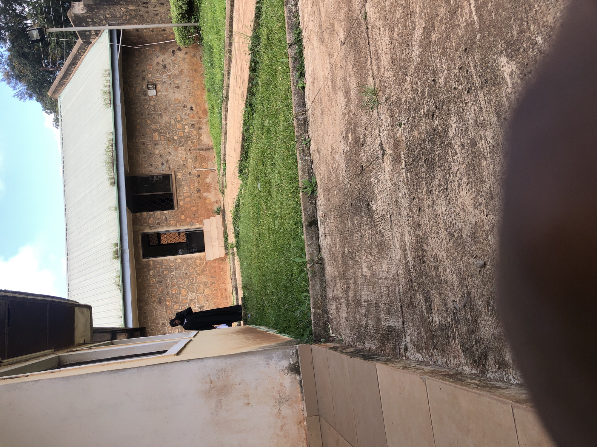 The image shows a stone building with a metal roof in Vom, Nigeria. A person wearing a long black garment and a headscarf stands near the building. The area appears to be an outdoor space with grass, concrete pathways, and some vegetation. The sky is visible in the background with scattered clouds. There is a light fixture on a pole to the right of the building.