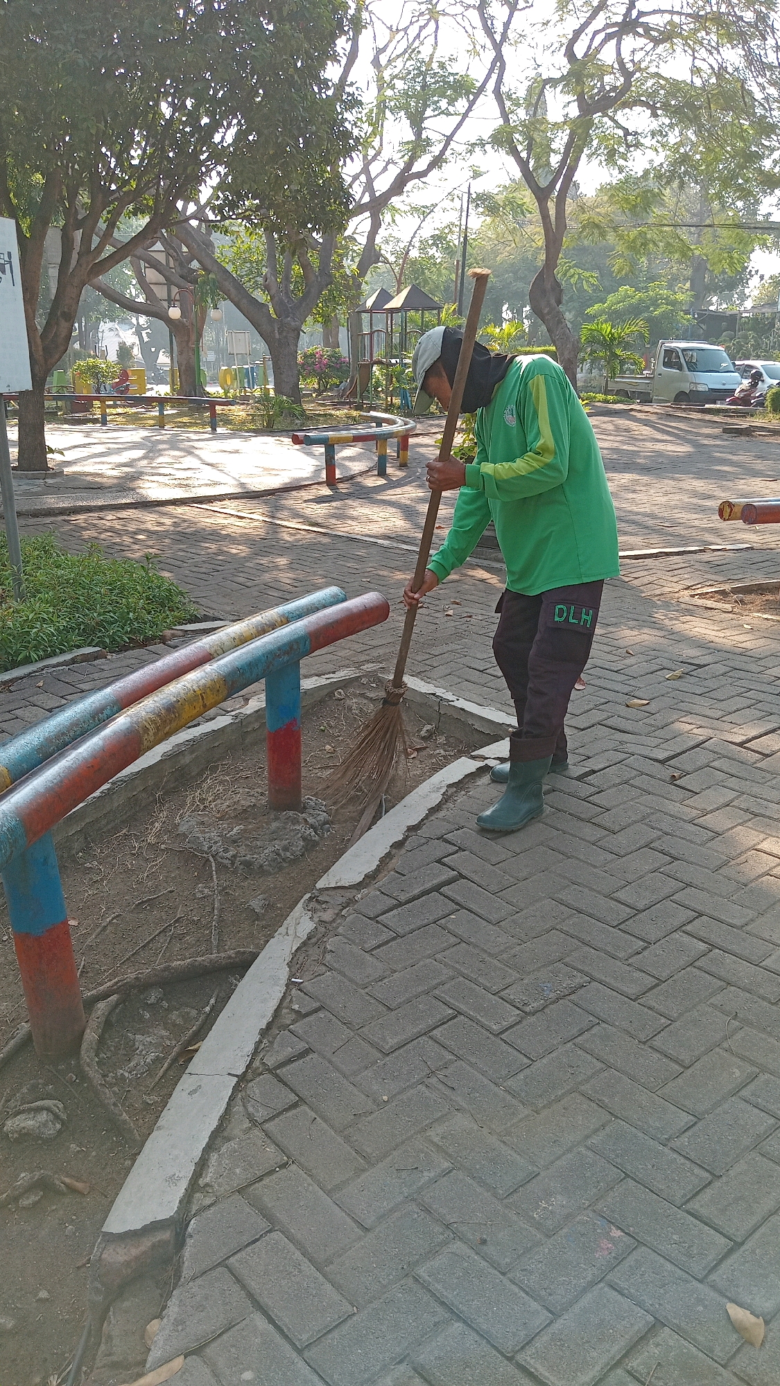A person wearing a green long-sleeved shirt and dark pants with "DLH" on the left leg is sweeping a paved area with a broom. The person is also wearing a cap and green boots. The scene appears to be in a park or outdoor public space with trees in the background, a playground structure, and benches. The ground is paved with brick-like stones in a herringbone pattern. There are also some metal poles with red, blue, and yellow paint on them. The time of day seems to be morning or late afternoon due to the shadows cast by the trees. The location is identified as Randuagung, Indonesia.