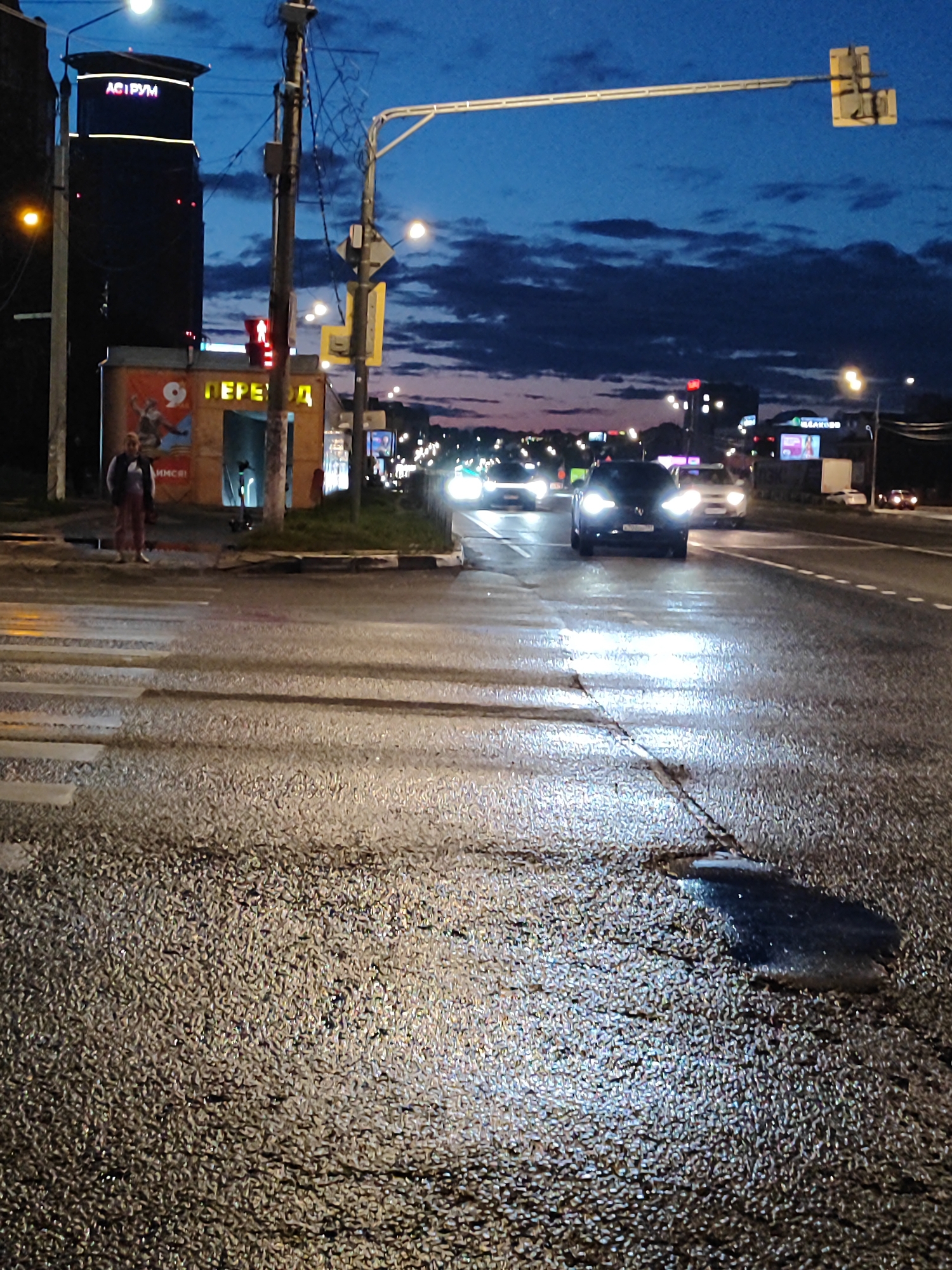 An evening shot captures a wet asphalt road at a pedestrian crossing in an urban environment. A dark, tall building with illuminated signage, "ACI PVM," stands prominently to the left. To the right of the building, a small kiosk with an orange awning displays the word "ПЕРЕХОД," meaning "Crossing." Several cars are driving on the road, their headlights reflecting on the wet surface. In the foreground, the pedestrian crossing itself is wet and reflective. A person stands on the sidewalk to the left, near the kiosk. The sky above is a deep blue with scattered clouds, indicating twilight or early night. Traffic lights and streetlights are visible, adding to the urban nighttime atmosphere.
