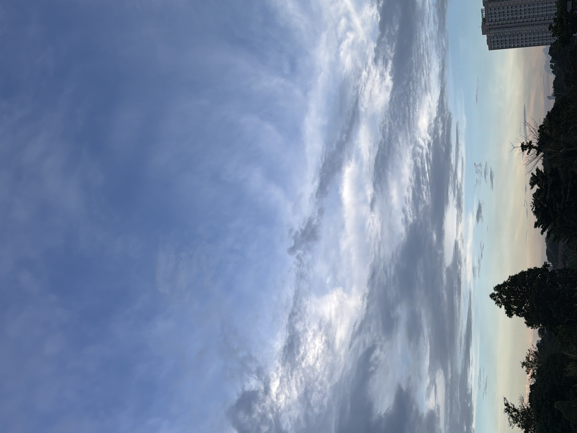 The image captures a cloudy sky over Davao City, Philippines, likely during the late afternoon or early morning. The sky is dominated by various cloud formations, ranging from wispy cirrus clouds to denser, darker stratus clouds. The clouds are illuminated by the sun, which is not directly visible, creating a soft, diffused light.
In the lower portion of the image, a dense line of trees forms a silhouette against the lighter sky. A prominent, tall building with many windows stands to the right of the frame, its architecture suggesting an urban environment. The building's height and design indicate it's likely an apartment complex or office building. The overall impression is one of a peaceful, natural landscape meeting the edge of a developed urban area. The image is composed to emphasize the sky, with the land and buildings serving as a grounding element at the bottom.