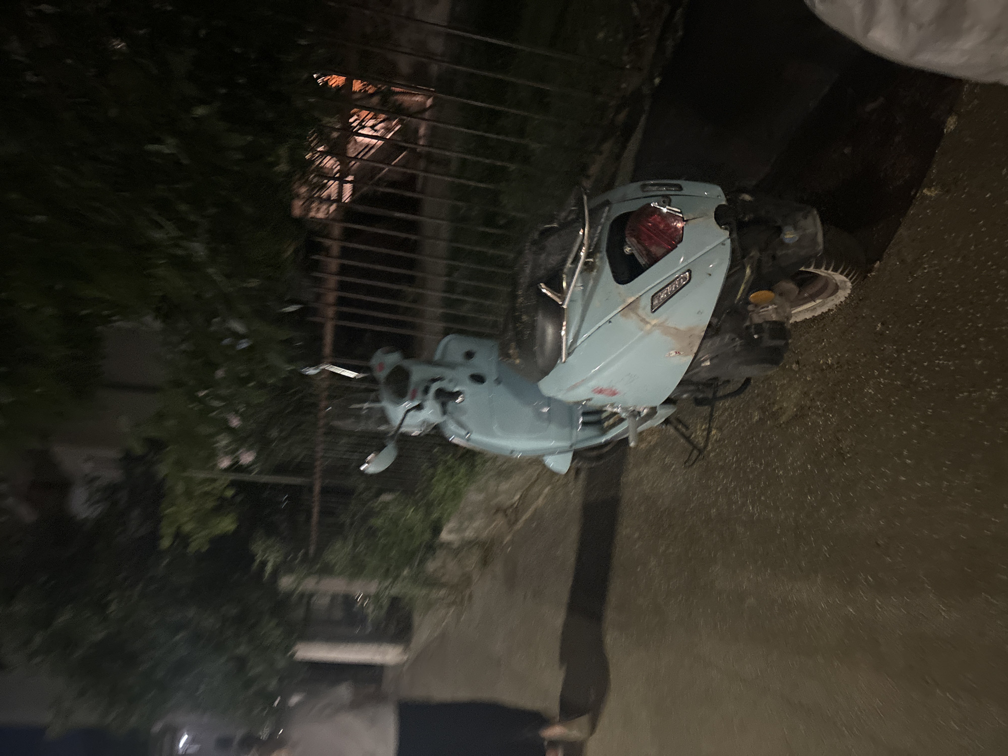 A pale blue scooter is parked on a paved street at night. The scooter is the primary focus of the image, with its rear end facing the camera. It appears to be slightly dusty or worn. Behind the scooter, a dark metal fence runs horizontally across the frame, separating it from a dimly lit area with trees and some foliage. The street surface is dark and wet, with scattered debris, possibly leaves or small stones. To the left of the scooter, a person is walking away from the camera, their form partially obscured by shadows. They are wearing dark pants and a light-colored top. The overall lighting suggests it is nighttime, with artificial light sources present but not strongly illuminating the scene. The context of Budva, Montenegro, suggests an urban or semi-urban environment typical of the region.