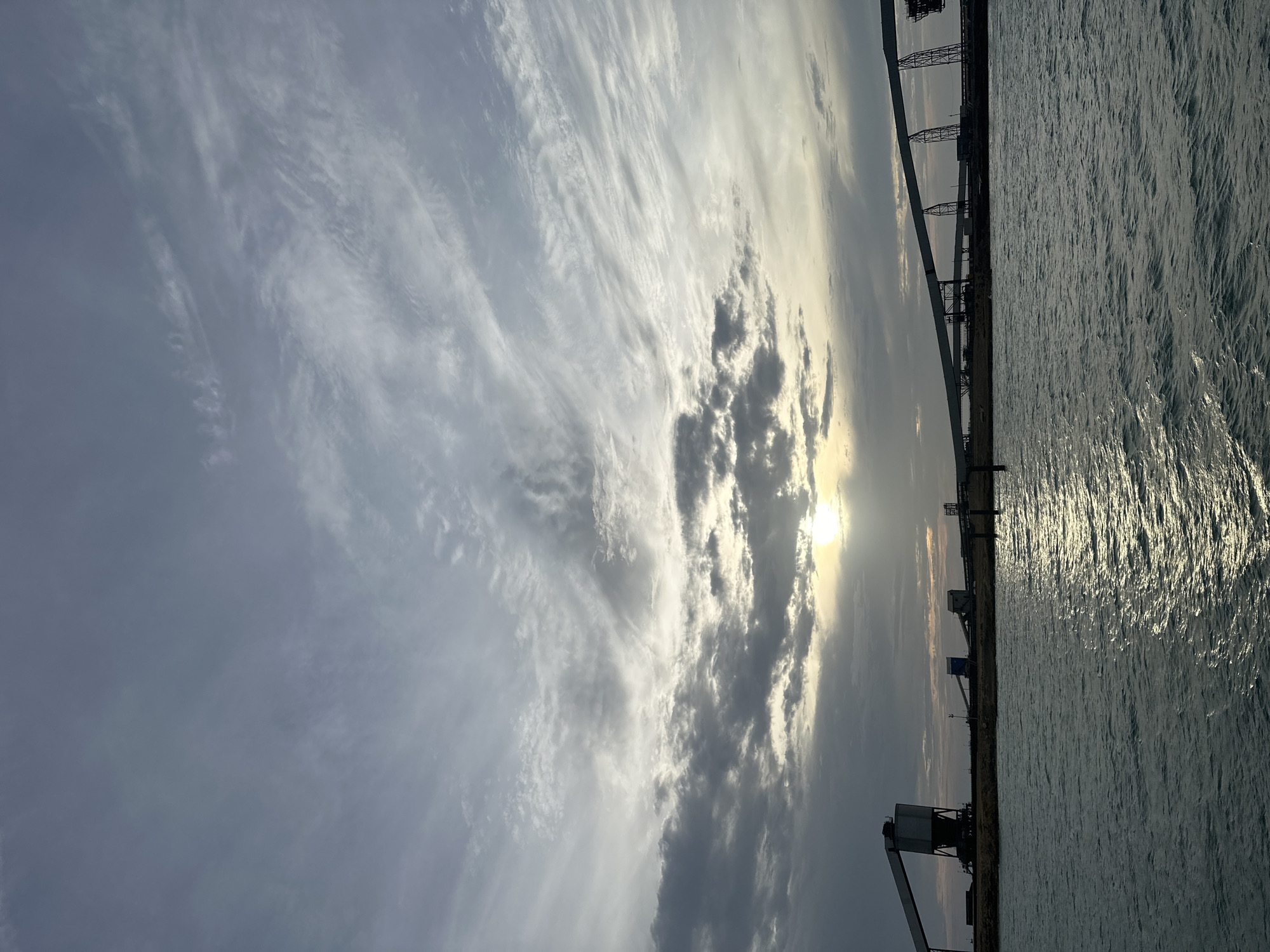 The image displays a wide-angle view of an industrial port facility at either sunrise or sunset, located in Uribia, Colombia. The sky occupies the upper two-thirds of the frame, characterized by a mix of stratocumulus and cirrus clouds, exhibiting various shades of grey and pale blue. The sun, a bright yellow-white disc, is partially obscured by clouds in the lower central sky, casting a strong reflection across the water in the foreground. The water, likely a sea or large bay, is dark blue-grey with distinct ripples and waves, showing a shimmering path of sunlight extending from the horizon towards the viewer.
On the flat horizon, silhouetted against the light, are multiple large industrial structures consistent with a bulk material handling port. On the left, a prominent tall loading terminal or silo with a large, angular conveyor belt system extends. Towards the center and right, an extensive series of elevated conveyor belts supported by multiple pylons stretches across the landscape, indicating a large-scale transport infrastructure. Other smaller industrial buildings, one with a distinct blue-grey color, are also visible between the larger structures. Two dark, slender vertical posts or markers are situated in the water in the mid-distance, closer to the shore. No people or active vehicles are present in the scene. The overall impression is one of an operational industrial port at a transition point of day, with the structures largely dark against the bright sky and reflective water.