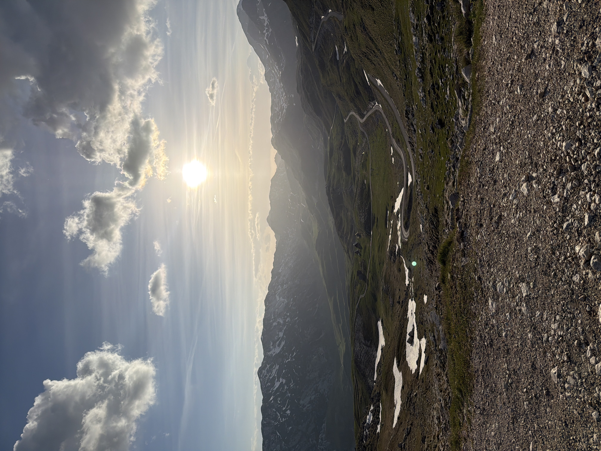 The image shows a mountainous landscape in Šavnik, Montenegro, bathed in sunlight. The sun is high in the sky, obscured by clouds. The mountains are covered in vegetation, with patches of snow still visible. A winding road snakes its way up the mountain. A rocky foreground completes the scene. There are no people visible.