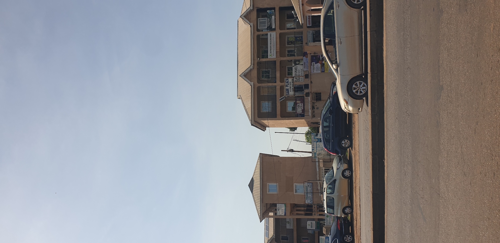 The image shows a street scene in Barnawa, Nigeria, featuring several commercial buildings and parked cars. The buildings are two or three stories high, with tan-colored exteriors and light brown, sloped roofs. The buildings have balconies with dark railings on the upper floors and windows. Several signs are visible on the building facades, including "SABR PLAZA," "OMEGA," and "ULTIMATE HEALTH HMO." Solar panels are installed on the roof of one of the buildings on the left.
Parked along the street are at least five vehicles. From left to right, these include a dark-colored sedan, a light-colored van, a dark blue SUV, and a gold sedan in the foreground. The road appears to be asphalt, with a concrete divider separating the lanes of traffic from the parking area. The sky is clear and bright blue. The overall scene depicts a typical urban environment with commercial activity.
