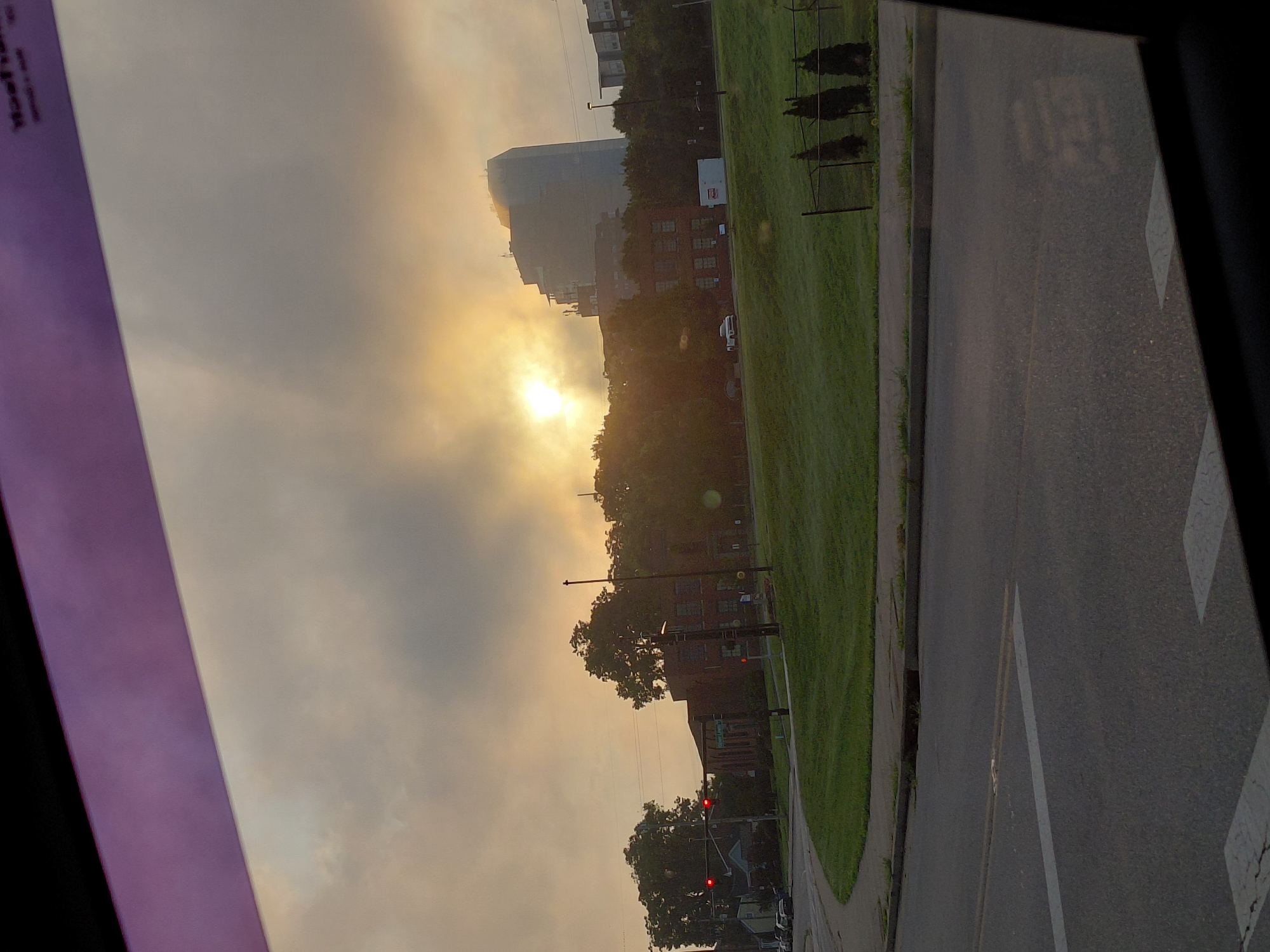 The image captures a hazy, sunlit scene from inside a vehicle, likely in Lexington, Kentucky, given the architectural style of the prominent glass building which resembles the Riverview Building at 200 West Vine Street in Lexington. The sun is breaking through a layer of clouds, creating a soft, diffused light that illuminates the sky and casts a glow on the surrounding environment.
In the foreground, a portion of a street and a sidewalk are visible, suggesting the vantage point is from a moving vehicle or a parked one at an intersection. The road is paved asphalt, with white lane markings. Beyond the road lies a grassy area, well-maintained and sloping gently upwards. A black chain-link fence runs along the edge of the grass, with three conical evergreen trees planted behind it.
Further back, a cluster of trees partially obscures a brick building with multiple windows. Across the street from this building, and slightly to the right, a striking, modern glass skyscraper stands tall, its curved facade reflecting the hazy sky. The building's unique shape is a key identifier. Smaller buildings and trees are interspersed in the middle ground, contributing to the urban landscape.
The overall atmosphere is one of early morning or late afternoon, with the diffused sunlight creating a serene, somewhat muted color palette. The presence of traffic lights, one of which is red, indicates an active urban environment. The purple tinted edge at the top of the image suggests the view is through a car's windshield or a tinted window.