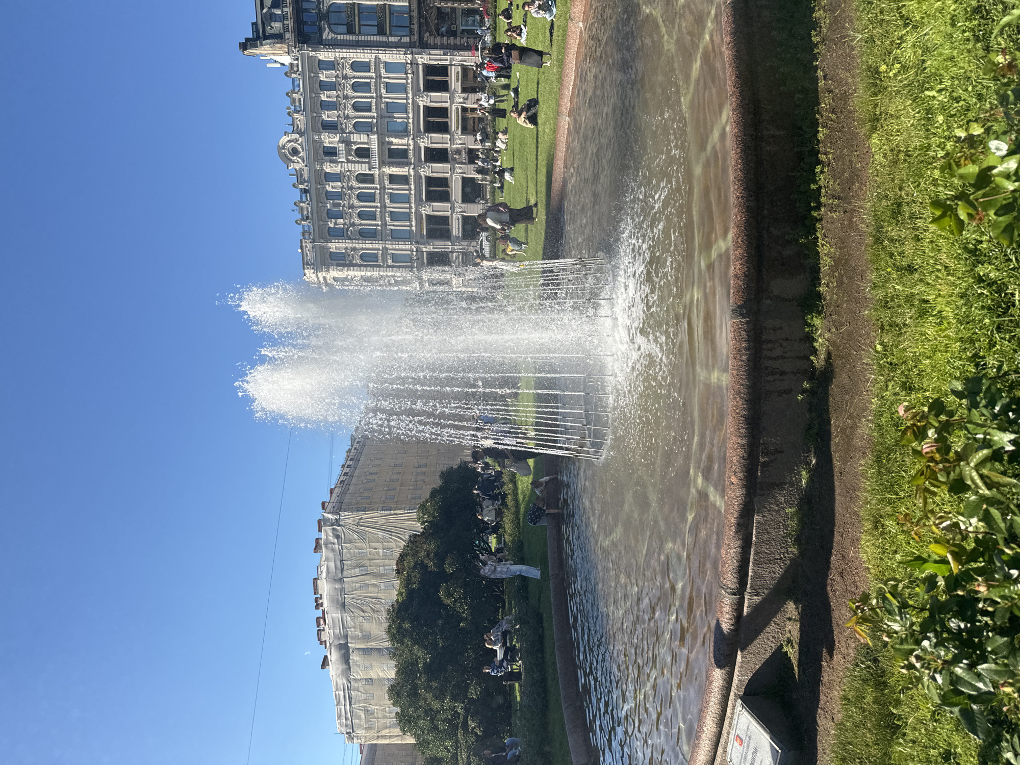 The image depicts a fountain in a city square in Saint Petersburg, Russia, under a clear blue sky. The fountain is in the foreground, with water spraying upwards. People are scattered across a grassy area in the background, some sitting, some standing, and some appearing to be interacting with the water from the fountain. Several buildings line the square, with one on the left covered in scaffolding. The architecture of the buildings suggests a historical European city. The overall scene is one of public recreation in an urban park setting.