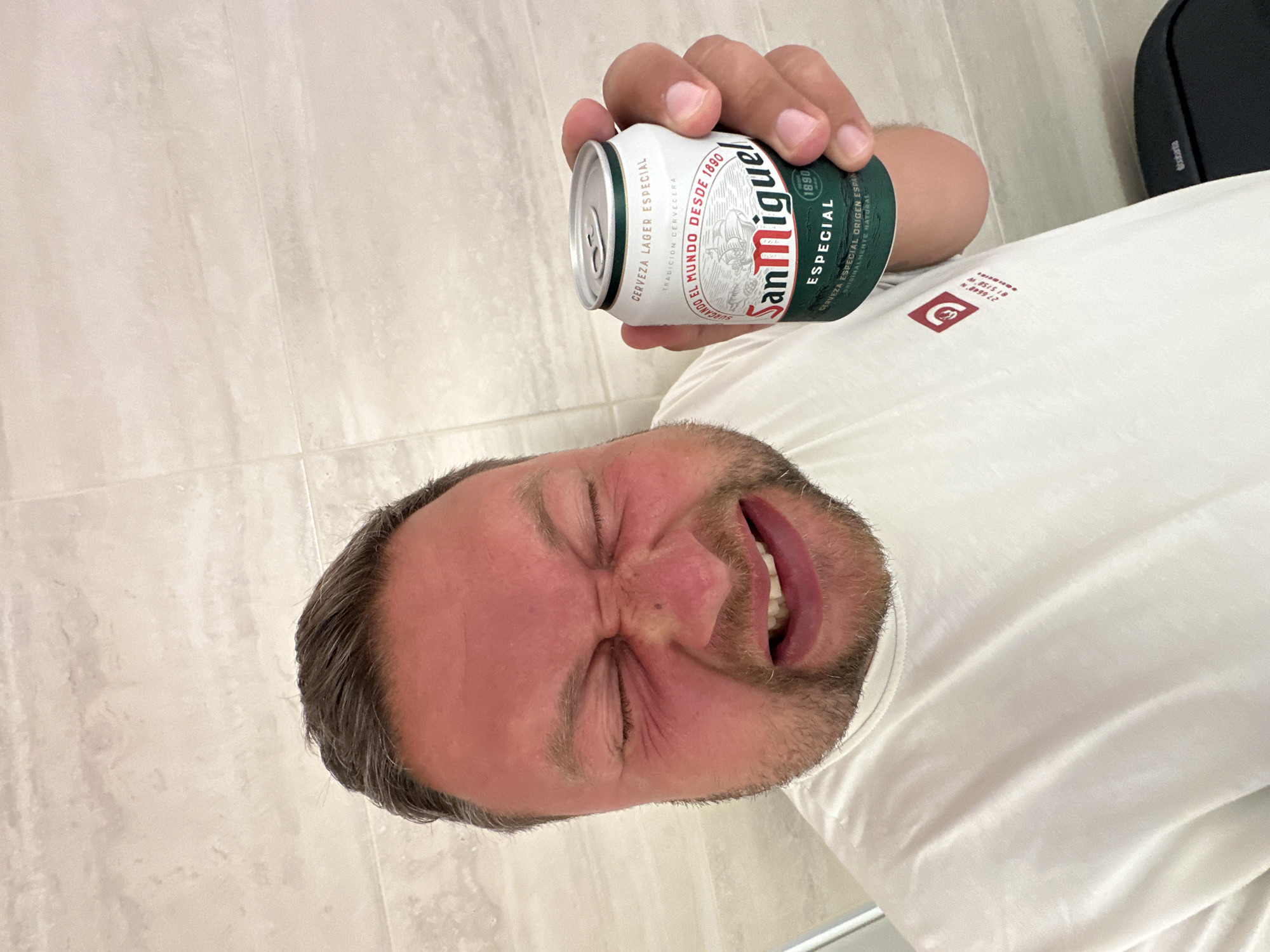 A man is holding a can of San Miguel beer and making a grimacing face in what appears to be a bathroom with tiled walls. The can is white and green with the San Miguel logo and text in Spanish. The man has short, graying hair and a beard, and he is wearing a white t-shirt. The background is a light-colored tile wall. The man is squinting his eyes and his mouth is open in a distorted expression. The image appears to be a selfie taken from a low angle.