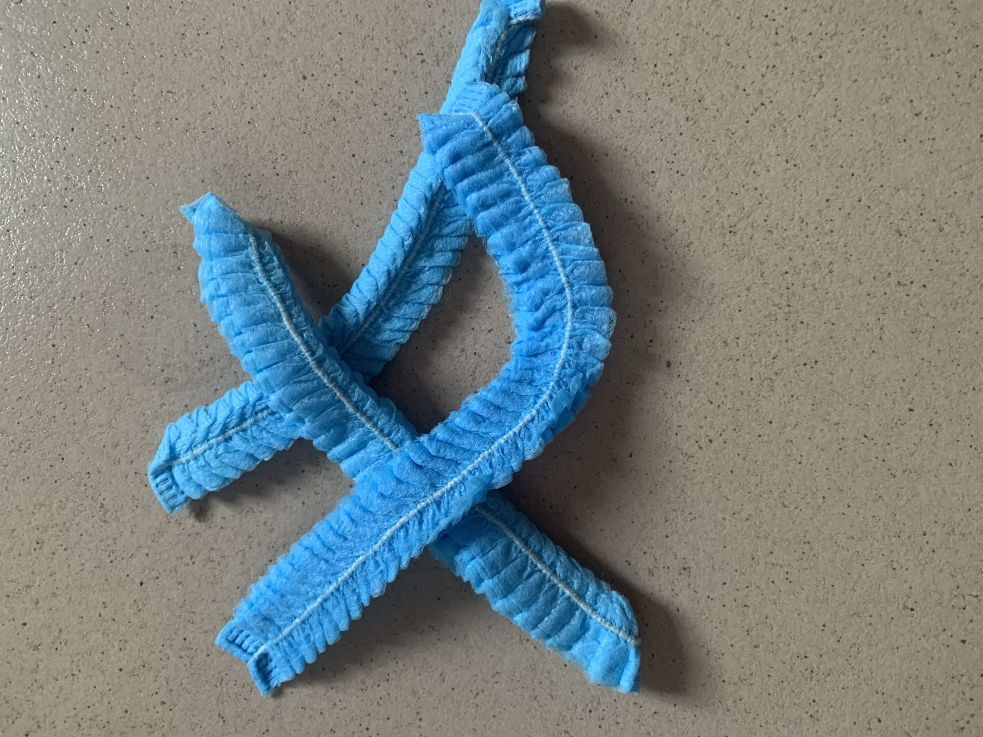 Two blue disposable hairnets are laid out in an "X" shape on a speckled grey surface. The hairnets are made of a crinkled, non-woven material and have a white elastic band around the open end. The surface appears to be a countertop or floor.