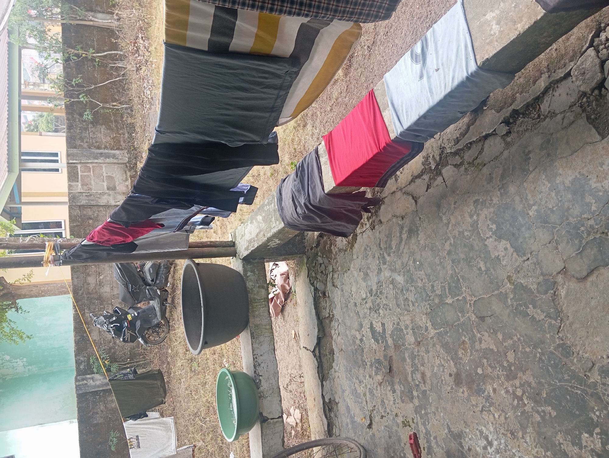 This image depicts an outdoor scene in Rejosari, Indonesia, likely a residential area given the presence of a home in the background. The foreground features a cracked concrete patio where laundry is drying. Several garments hang from a clothesline strung between two poles, including t-shirts, pants, and striped fabric. Below the clothesline, a concrete ledge displays more folded laundry, including a red towel and a light blue t-shirt. Two large basins, one green and one dark gray, sit on the ledge. Behind the basins, a black motorcycle is parked. The background shows a low concrete wall, dry vegetation, and a light-colored building with a green roof. The overall impression is a simple, practical setup for laundry in a warm climate.