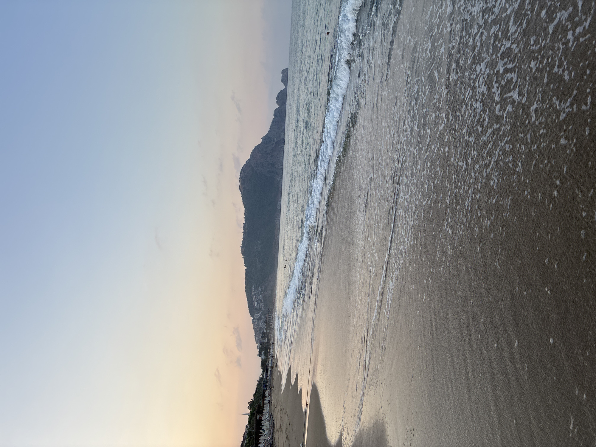 The image depicts a tranquil beach scene at dawn in Alanya, Turkey. The sky transitions from a pale yellow and pink near the horizon to a soft blue overhead. A gentle wave washes onto the sandy shore, leaving a thin layer of foam. In the distance, a mountainous peninsula with scattered buildings is visible, characteristic of Alanya's landscape. The beach is lined with rows of white lounge chairs and umbrellas, suggesting a resort area. The water is calm with small waves, and a few individuals can be seen swimming or wading in the sea. The overall impression is one of peacefulness and the start of a new day.