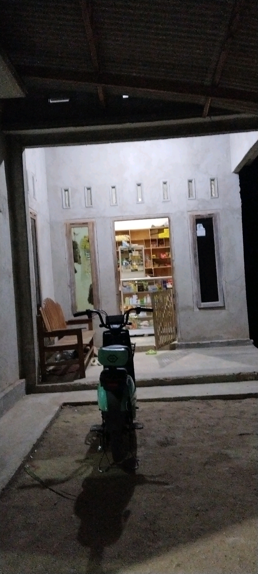 This image depicts the exterior of a small shop or convenience store at night in Tanjung Ratu Ilir, Indonesia. The building has a light-colored concrete facade with multiple horizontal, rectangular openings near the top of the entrance. The entrance itself is divided into three sections: a door on the left, an open doorway leading into the shop in the center, and a closed door on the right. Through the central open doorway, shelves stocked with various items, likely merchandise, are visible. A dark-colored motorbike is parked directly in front of the central entrance, casting a long shadow on the dirt ground. To the left of the motorbike and the entrance, there is a wooden bench with a high back. The ground in front of the building is covered with dirt and small stones. Above the building, a corrugated metal roof structure is visible. The overall lighting suggests it is nighttime, with artificial light emanating from within the shop.