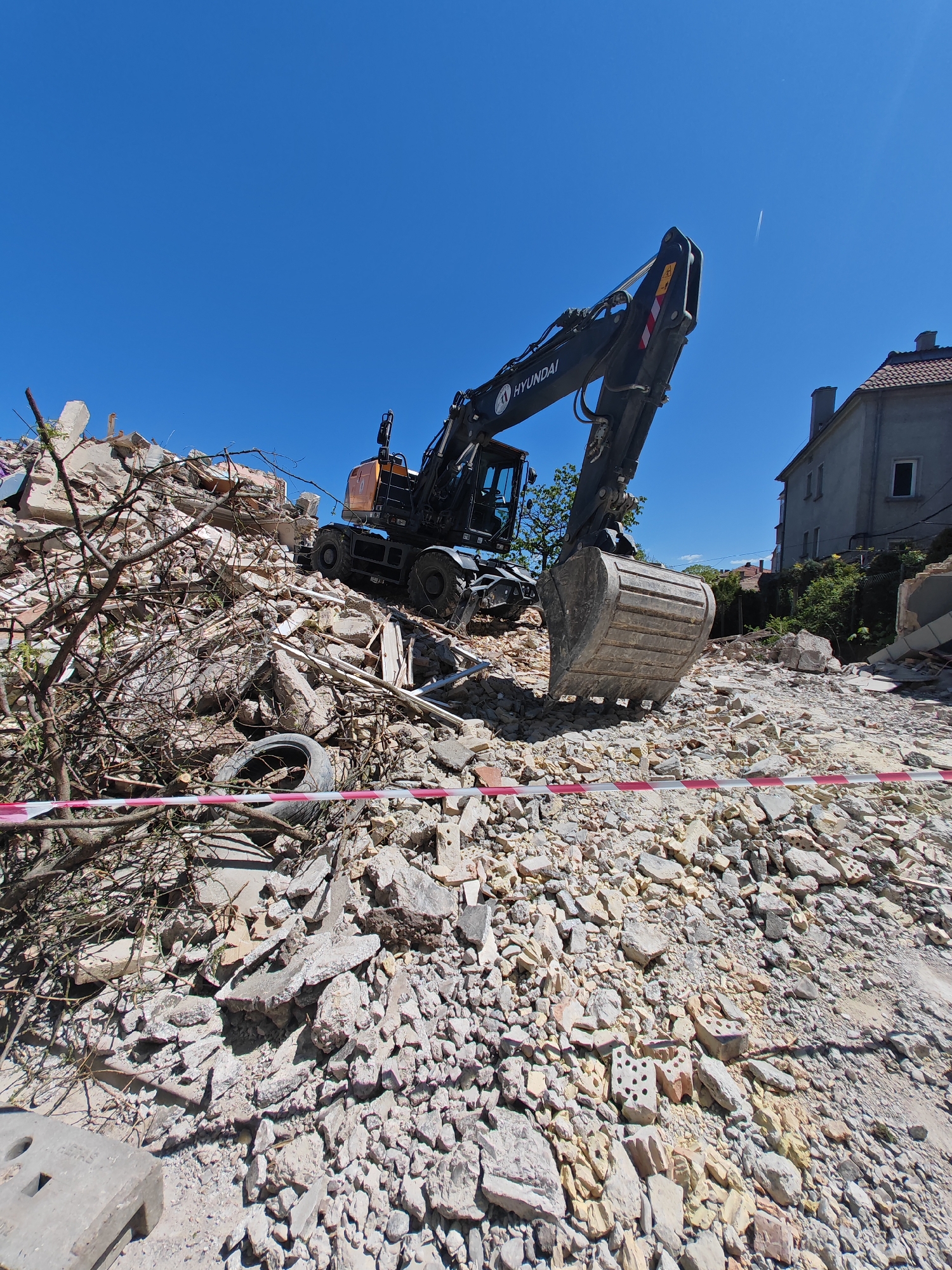 The image shows a demolition site, likely in Shumen, Bulgaria, featuring a Hyundai excavator positioned atop a mound of rubble. The debris consists of broken concrete, bricks, and various construction materials. A discarded tire is visible within the rubble pile, and red and white striped barrier tape stretches across the foreground. A building can be seen in the background. The sky is clear and blue.