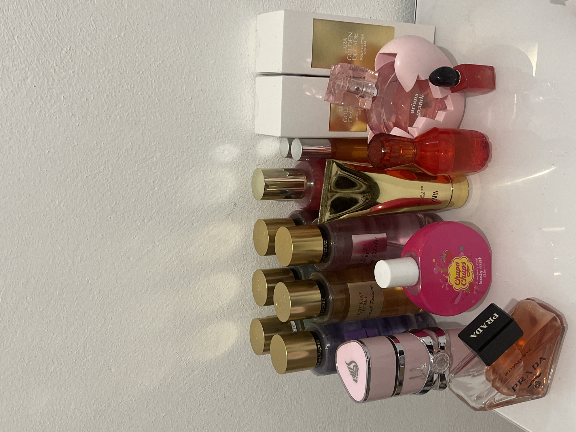 The image shows a collection of various perfume bottles and body sprays arranged on a white surface against a plain white wall. The collection includes multiple Victoria's Secret body mists with gold caps, a pink Prada perfume bottle, a Chupa Chups body mist, a red-orange perfume bottle, a pink Ariana Grande perfume bottle, two Zara Golden Decade body glitter cream boxes, a gold tube, and a small red perfume bottle with a black cap. The scene is indoors, and the lighting suggests it is daytime, possibly with natural light casting shadows from the left. There are no people or animals visible in the image. The arrangement appears to be a still life composition on a shelf or table. The location in Berlin, Germany, cannot be determined from the visual information provided.