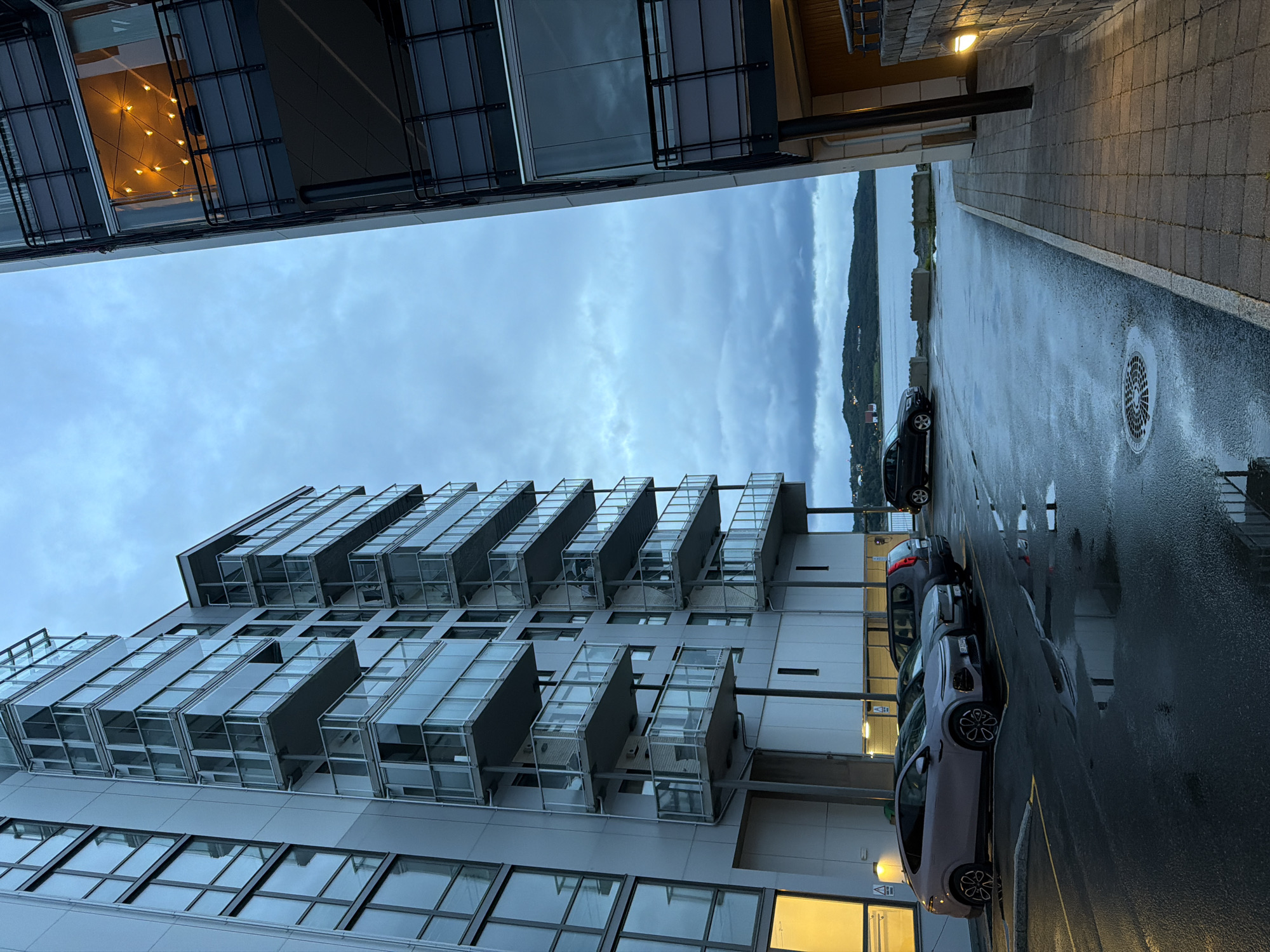 This is a vertical, eye-level, outdoor, full shot image of modern apartment buildings with balconies and windows, a parking lot with several cars, and a body of water in the background. The lighting suggests it is dusk or early evening, with a cloudy sky. The apartment buildings appear to be made of concrete and glass, with contemporary architectural designs. One building has a section of windows lit from within, revealing decorative string lights. The parking lot is wet, indicating recent rain, with puddles reflecting the surrounding environment. In the background, a body of water is visible, with a low, green, hilly landscape on the far shore, dotted with houses and trees. The image has been identified as being taken in the city of Harstad, Norway.