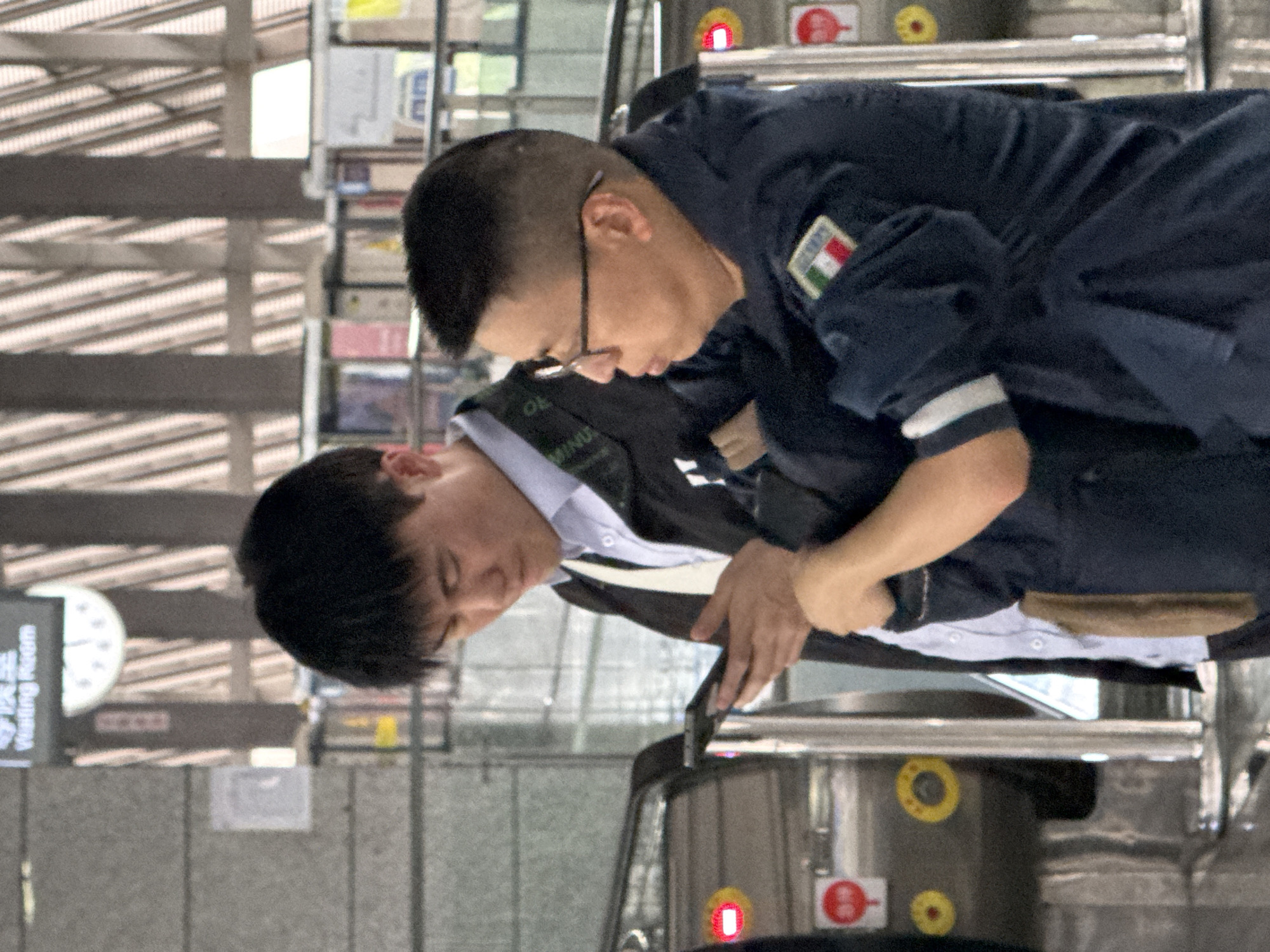 The image shows two Asian men standing in an indoor location. One man, wearing glasses and a dark blue uniform with what appears to be an Italian flag patch on the sleeve, is leaning over. The other man is wearing a white shirt, a dark jacket and holding a mobile device. The surroundings appear to be a public space, possibly a waiting area or transportation terminal with signs written in Chinese visible in the background.