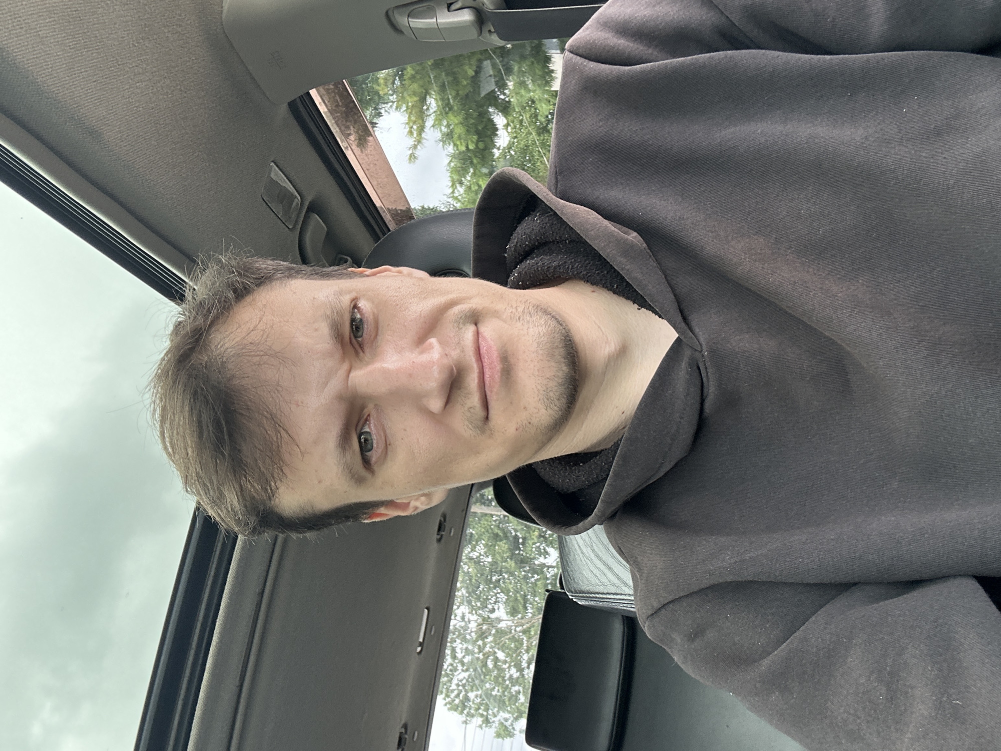 A man is in the driver's seat of a car. He is wearing a dark gray hooded sweatshirt. The sunroof of the car is open, revealing an overcast sky. Trees are visible through the car windows. The man is looking at the camera with a slightly furrowed brow. His hair is brown and short, with some thinning on top. He has a short beard and stubble. The background is blurred, but appears to be a residential area. The lighting suggests it is daytime. The overall atmosphere is casual and somewhat mundane.
