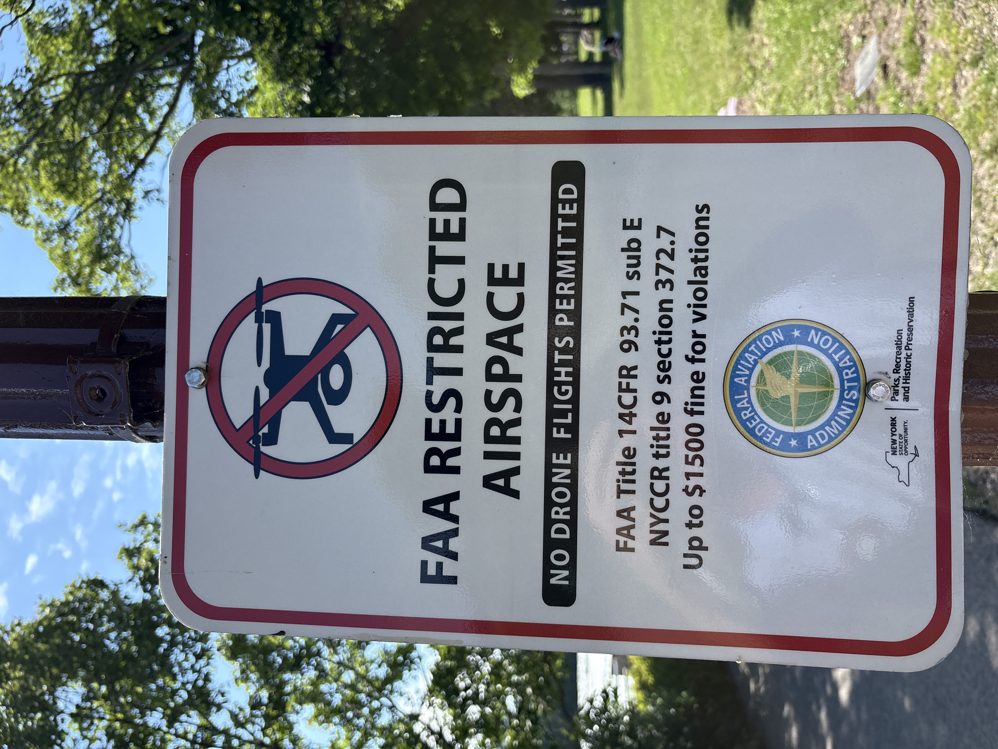 The image is a close-up shot of a white sign with a red border, mounted on a dark pole. The sign is oriented vertically and displays text and graphics indicating restrictions on drone flights. At the top, there is a circular symbol with a drone inside, crossed out with a red line, signifying "no drones." Below this, large black text reads "FAA RESTRICTED AIRSPACE." Further down, a black rectangular bar contains white text: "NO DRONE FLIGHTS PERMITTED." Beneath this, smaller black text provides legal citations: "FAA Title 14CFR 93.71 sub E" and "NYCCR title 9 section 372.7," followed by "Up to $1500 fine for violations." At the bottom of the sign, there are two logos: one circular logo with "FEDERAL AVIATION ADMINISTRATION" around it, and to the left, the outline of New York State with the text "NEW YORK STATE OF OPPORTUNITY." To the right of the New York logo, there is a smaller logo with "Parks, Recreation and Historic Preservation."
The background consists of a blurred green grassy area with trees and a blue sky with some clouds. The lighting suggests it is daytime. There are no discernible people or specific actions occurring in the immediate foreground, but in the distant background, a person is visible sitting on the grass. The overall scene appears to be an outdoor public space, likely a park, where drone usage is prohibited due to airspace regulations. The specific location within the city of Niagara Falls, United States of America, cannot be determined from the visual information presented.