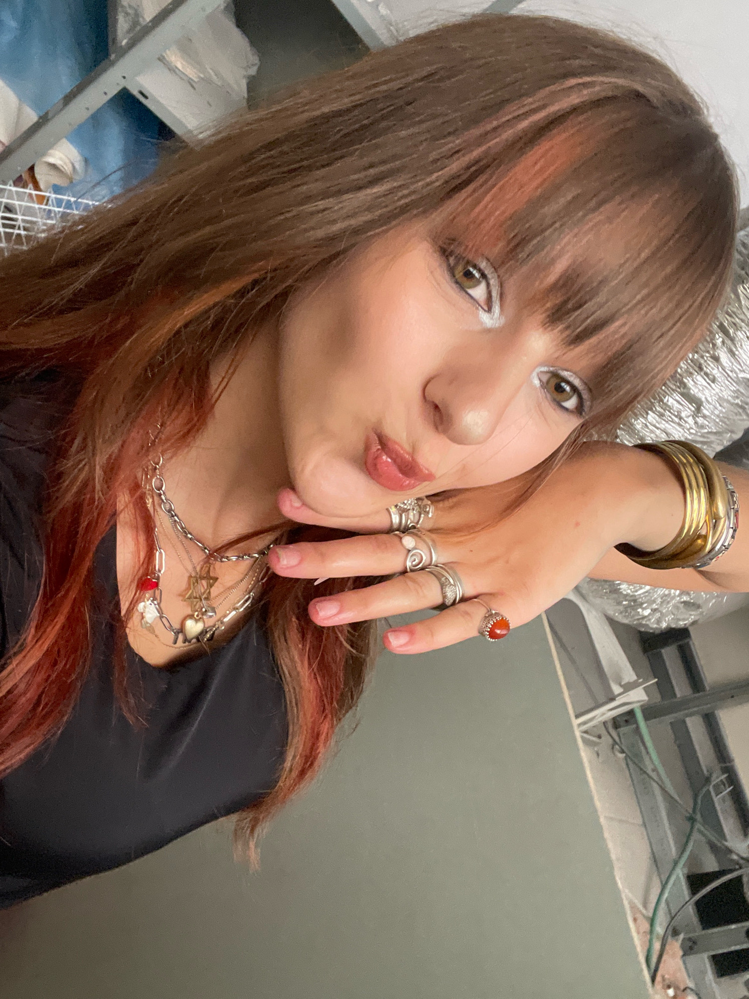 The image is a close-up, high-angle selfie of a young woman with medium-length, brown hair with reddish-orange highlights and bangs. She is wearing silver eyeshadow and has her mouth puckered. She is wearing multiple layered necklaces with various charms, including a red bead, a white figure, a Star of David, and a heart. She also has several rings on her fingers, including one with an orange stone. On her right wrist, she is wearing several gold-colored bangles. She is wearing a black top. The background appears to be indoors, with some exposed shelving and wiring, suggesting a construction or unfinished space. The image does not contain any information that would allow it to be located in Raanana, Israel.