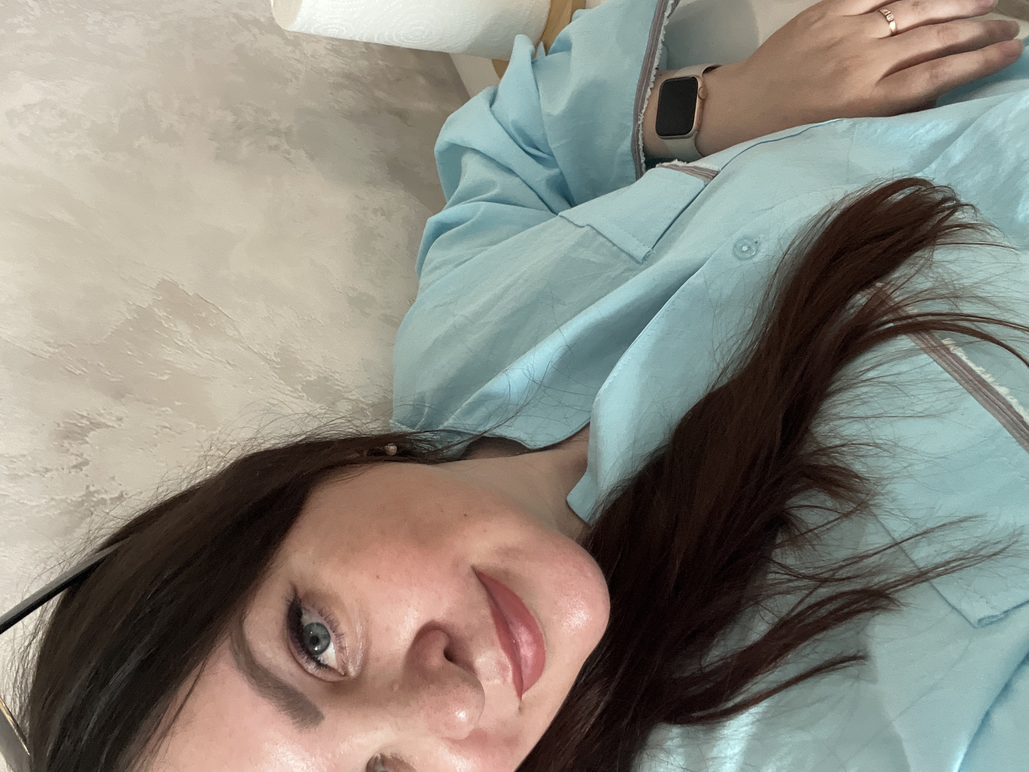 A woman is lying on a light-colored, possibly textured surface. She has dark brown hair, fair skin, and light-colored eyes. She is wearing a light blue button-down shirt. Her left arm is visible, and she is wearing a smartwatch and a ring on her finger. A partially visible roll of white paper is in the upper left corner of the image. The image is taken at an angle, looking down at the woman. The location in the city of Izhevsk, Russia, is not determinable from the image.