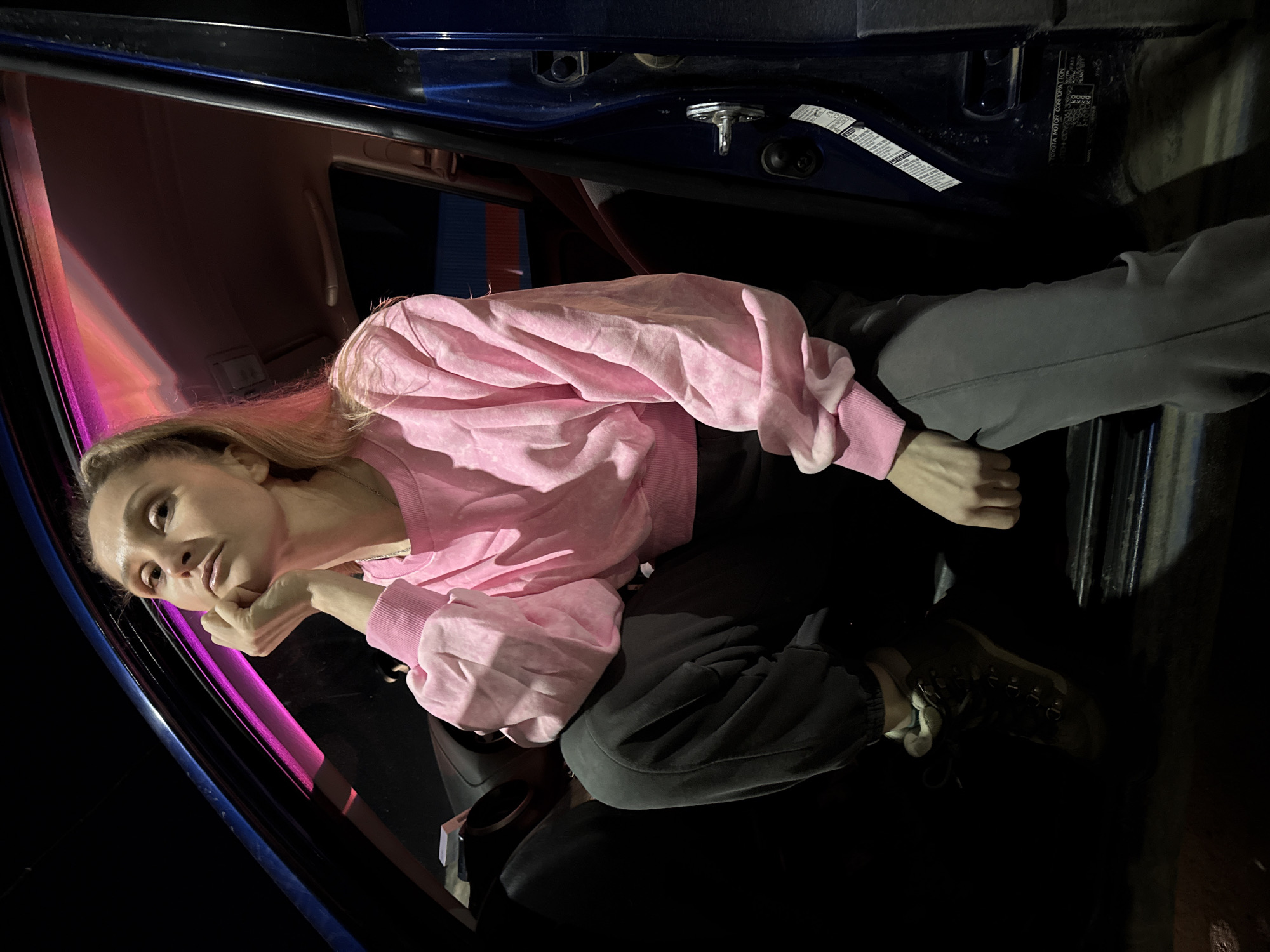 A woman sits inside a dark blue car. She wears a pink sweatshirt, dark pants, and hiking boots. The location is unknown, with no details suggesting Chaikovsky, Russia. The only source of light is a pink glow that could be from the interior of the car. The woman is resting her face on her hand and appears to be looking out the window. There is a blue door frame next to her with an informational sticker.