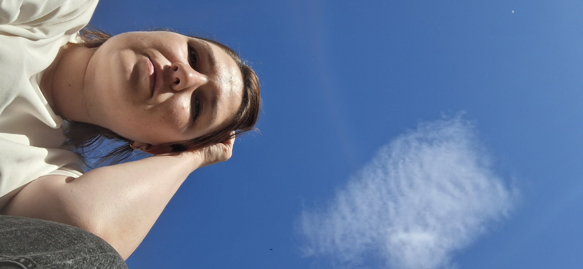 This media file is a low-angle, close-up shot of a person's face and upper body against a clear blue sky. The subject, a woman, is resting her head on her hand, looking upwards. She has brown hair, and her expression appears relaxed. She is wearing a white shirt. The sky is bright and cloudless except for a wispy cloud formation in the lower right portion of the frame. A tiny dark speck, possibly a bird, is visible in the sky below the cloud. The lighting suggests it is daytime, likely midday due to the brightness. There are no visible text elements in the image. The perspective and clear sky suggest an outdoor setting, potentially during fair weather.