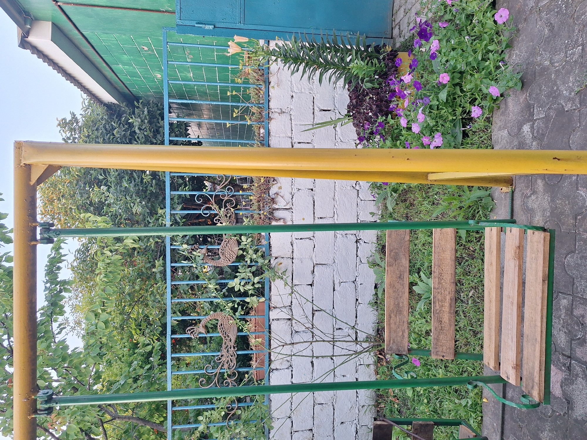 The image depicts an outdoor scene in Nabran, Azerbaijan, featuring a swing set in the foreground. The swing set has a yellow horizontal bar supported by green vertical posts, with a wooden seat consisting of several planks. Behind the swing set, a white brick wall is visible, topped by a blue metal fence. Decorative metal peacock figures are attached to the fence. Beyond the fence, lush green trees and foliage create a natural backdrop. To the right of the swing set, a vibrant flower bed filled with purple and pink petunias and other flowering plants adds a splash of color. A tall, slender green plant with long leaves stands prominently in the flower bed. In the far right of the image, a portion of a green building with a white trim roof is visible. The ground is paved with interlocking grey tiles. The lighting suggests it is daytime, possibly late afternoon or early evening, given the soft light and shadows. There are no people present in the image.