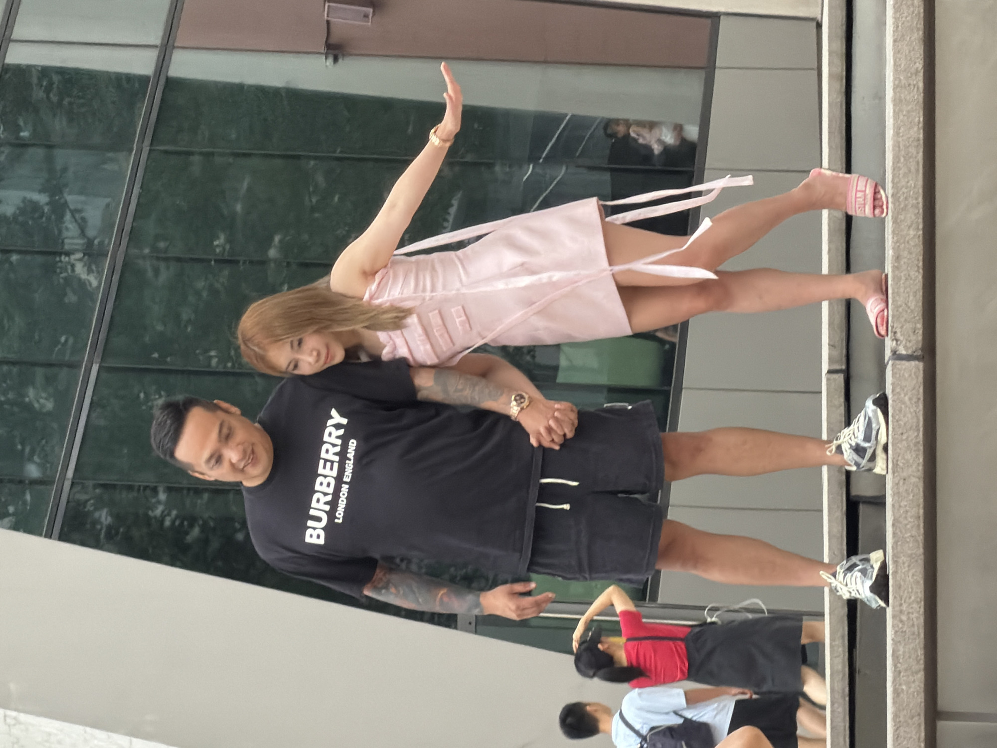 Two individuals are standing on a concrete ledge in front of a modern building with a glass facade. The person on the left is a man with tattoos on his arms, wearing a black t-shirt with the "BURBERRY LONDON ENGLAND" logo and dark shorts. He is holding hands with the person on the right, a woman with shoulder-length blonde hair. She is wearing a light pink, strapless dress with ribbon detailing and pink slides. The woman is extending her left arm outwards. In the background, several other people are visible, including a person wearing a red shirt and black skirt and another person in a blue t-shirt and backpack. The environment appears to be an urban setting in Yuzhong District, China, characterized by contemporary architecture.