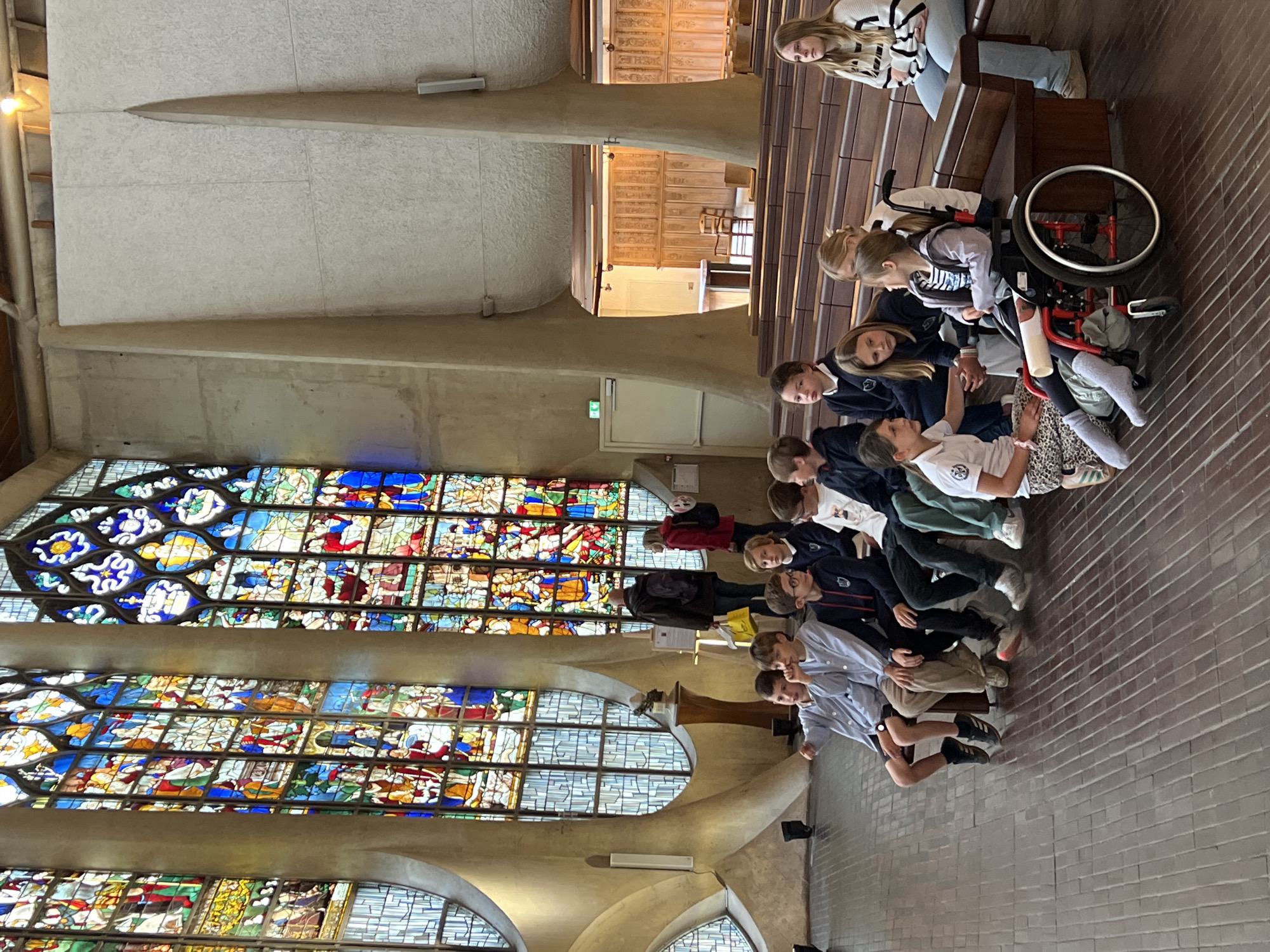 This image captures the interior of the Église Sainte-Jeanne-d'Arc in Rouen, France, characterized by its modernist concrete architecture and large, multi-paneled stained-glass windows. The floor is composed of rectangular, reddish-brown tiles laid in a stretcher bond pattern.
On the left, several tall, arched stained-glass windows, featuring both traditional figurative scenes in vibrant colors and more geometric, clear glass sections, are set into the concrete structure. Between two windows, a small white bust rests on a dark wooden pedestal. A green exit sign is visible on a concrete wall section further back on the left.
In the foreground and mid-ground, a group of approximately 15 school-aged children, mostly pre-teens, are present. Many are seated on tiered, dark wooden benches that arc across the right side of the frame. On the front-most bench to the left, two boys in light blue uniform shirts are seated; one points towards the left. Further to the right, a girl in a red wheelchair, wearing a leg brace on her right leg, is on the tiled floor. Another girl, wearing a white shirt and patterned leggings, sits on the floor beside the wheelchair, leaning in and interacting with the girl in the wheelchair. On the far right end of the benches, a woman in a white and black striped sweater and blue jeans is seated, looking towards the left.
In the background, on the left, two adults stand near the stained-glass windows. One adult, wearing a dark jacket, faces away from the camera while carrying a yellow bag. The other adult, in a red jacket, faces the first. Further back on the right, through an architectural opening, a section of dark wooden wall paneling and a simple wooden chair are visible.