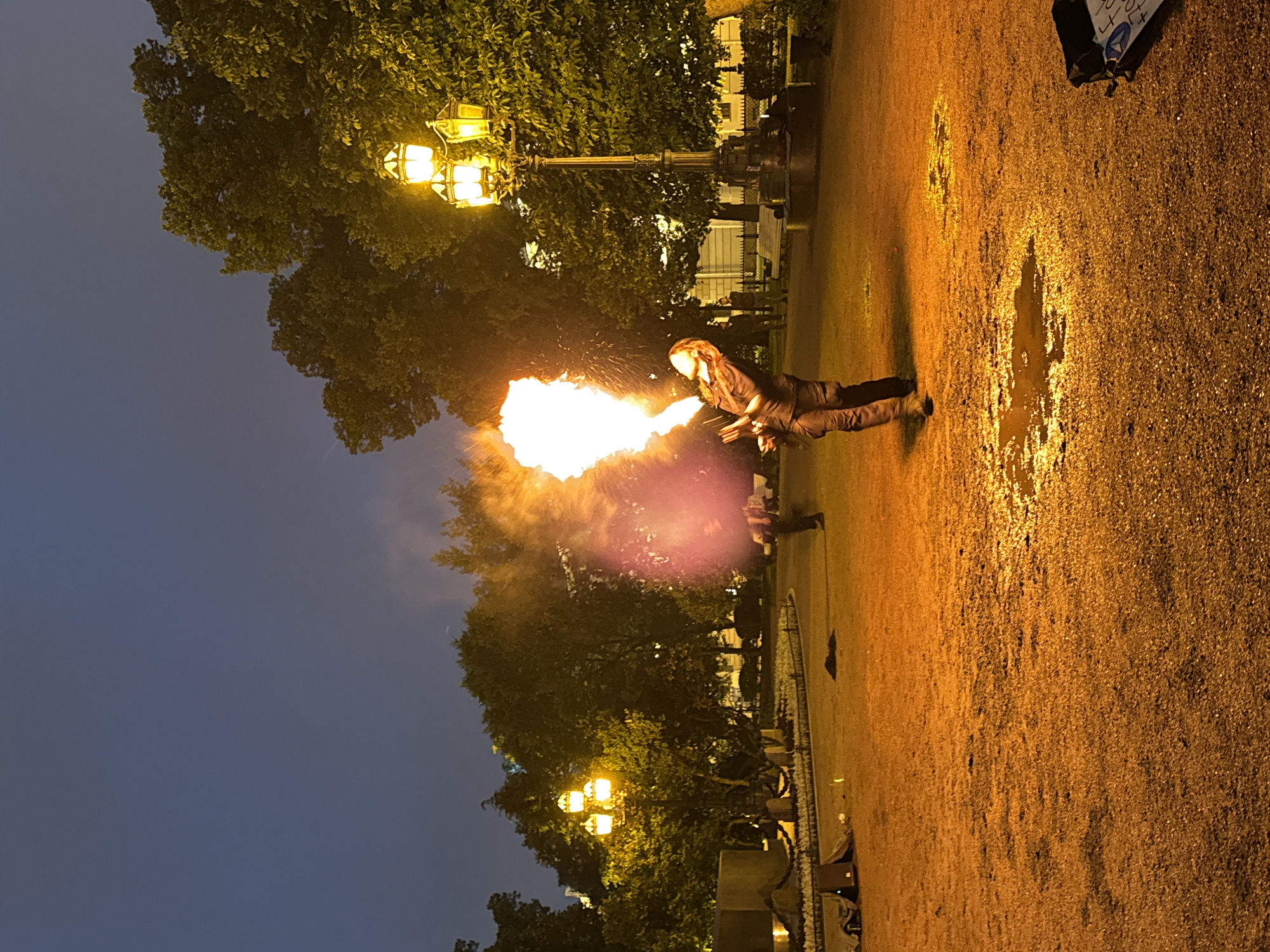 In Palace Square in Saint Petersburg, Russia, at dusk, a fire performer exhales a large plume of flame. The performer, dressed in dark clothing, stands in the center of the image, head tilted back as they direct the fire away from themselves. The flame illuminates the surrounding area with an orange glow, casting shadows. The ground, covered in gravel and small puddles of water reflecting the light, appears wet. Behind the performer, several trees with dense foliage are visible, and a vintage street lamp with three glowing orbs stands to the right, its light adding to the warm ambiance. In the background, other figures can be seen walking, and the silhouettes of buildings suggest an urban setting. The sky above is a deep blue, transitioning from twilight.