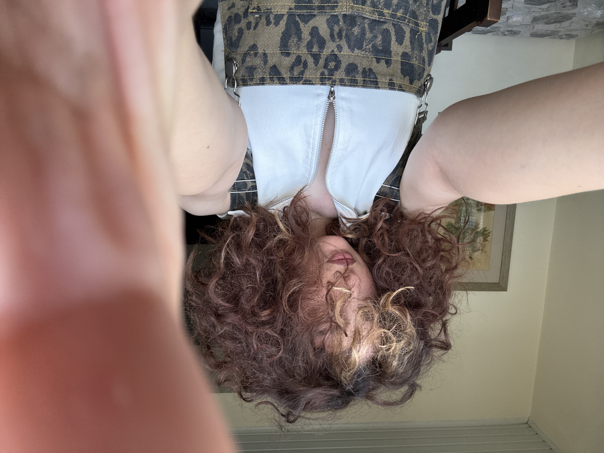 A person with brown curly hair is positioned upside down in the frame. They are wearing an outfit with a white top and brown animal print overalls. The background appears to be an interior setting, with light walls and a framed picture. The image does not provide enough detail to determine if the location is in Yerevan, Armenia.