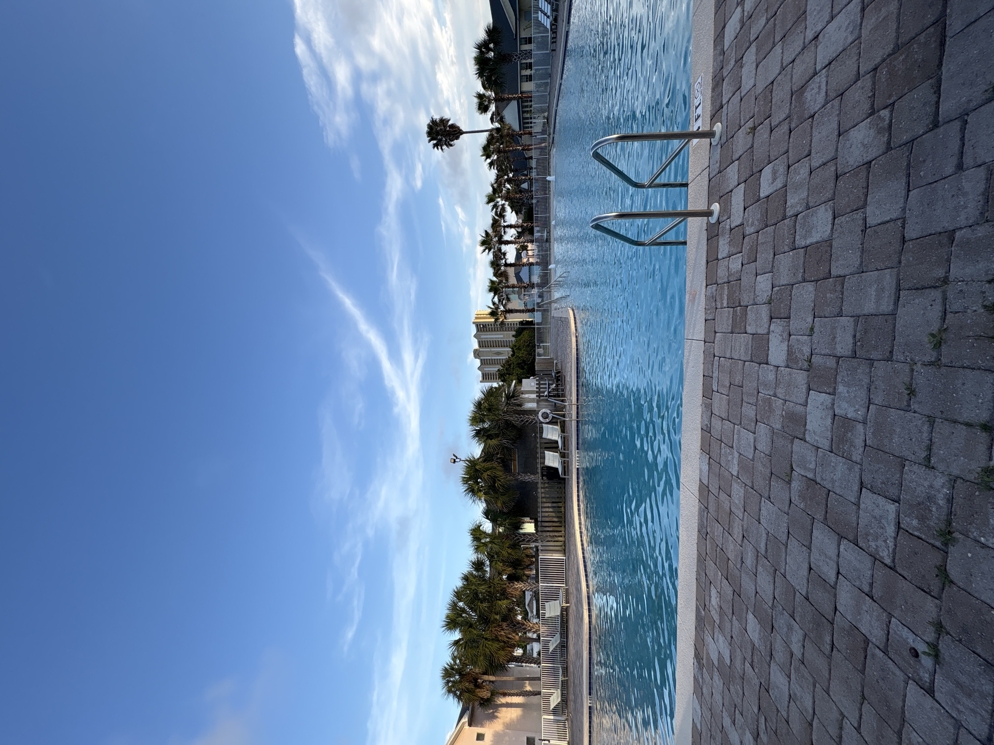 The image depicts a swimming pool area in Panama City Beach, Florida. A clear, blue sky with wispy clouds stretches above. The pool's water surface is rippled, reflecting the sky. Metal handrails lead into the pool. A paved area of interlocking bricks borders the pool. Beyond the pool, a line of palm trees and buildings can be seen. White lounge chairs are scattered along the pool deck, and a white fence separates the pool area from the surrounding landscape. In the distance, there are high-rise buildings.