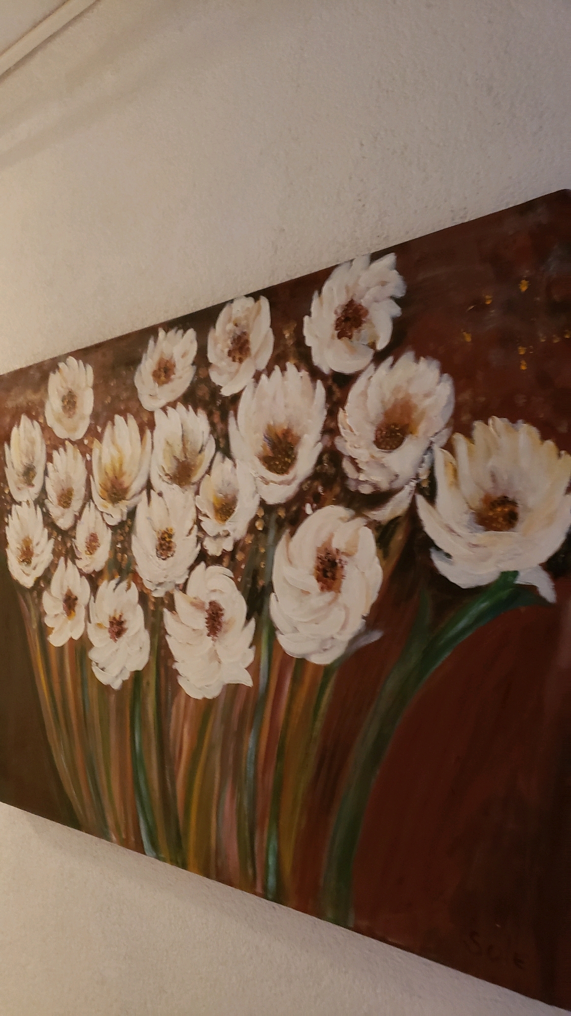 This is an image of a painting of white flowers with brown centers, displayed on a white wall. The painting is elongated horizontally and features a bouquet of these flowers against a dark brown background with gold flecks. The flowers vary in size and stage of bloom, with some appearing more open and others more bud-like. Thin, vertical brushstrokes in shades of green, yellow, red, and brown suggest stems and foliage. In the bottom right corner of the painting, a signature is visible, reading "S.C.E.". The painting is positioned in what appears to be an indoor setting, possibly a hotel room or private residence, given the textured white wall and the angle of the artwork. The location of the painting is not identifiable from the image itself, however, if it were located in Općina Tučepi, Croatia, it would be displayed within a building in that municipality on the Dalmatian coast.