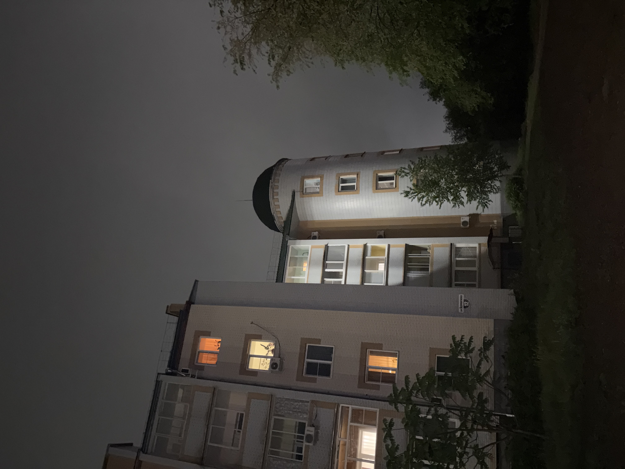 The image, captured at night, presents a view of two buildings under a dark, overcast sky. On the right, a distinctive tall, cylindrical building features a dark, rounded top and evenly spaced rectangular windows, some showing interior illumination. Its facade is light-colored with darker trim around the windows. To the left, a multi-story rectangular apartment building is visible, with many windows glowing yellow and white from interior lights, suggesting occupancy. Air conditioning units are installed below some windows. Dark tree branches with sparse foliage frame the upper and lower edges of the frame, partially obscuring the sky and parts of the buildings. On a light-colored section of the apartment building, a blue sign with white Cyrillic text clearly reads "ул. Бумагина 12" (Bumagina Street 12). The sky is uniformly dark grey, indicating a heavily clouded night. The scene depicts a quiet urban environment at night with no visible human activity or specific events taking place.