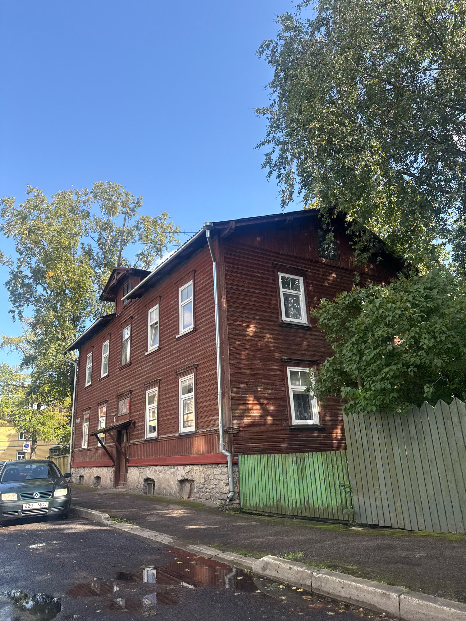 The image depicts a two-story wooden house in Tallinn, Estonia, with a dark red exterior and white-trimmed windows. The house has a pitched roof and a small dormer window on the upper level. A portion of a green wooden fence is visible in the foreground, bordering a paved sidewalk and a street. A dark green Volkswagen car is parked on the street to the left, and puddles of water are present on the asphalt. Tall trees with green foliage surround the house, casting shadows on its facade. The sky is clear and blue. The architectural style suggests an older building, common in residential areas of Tallinn.